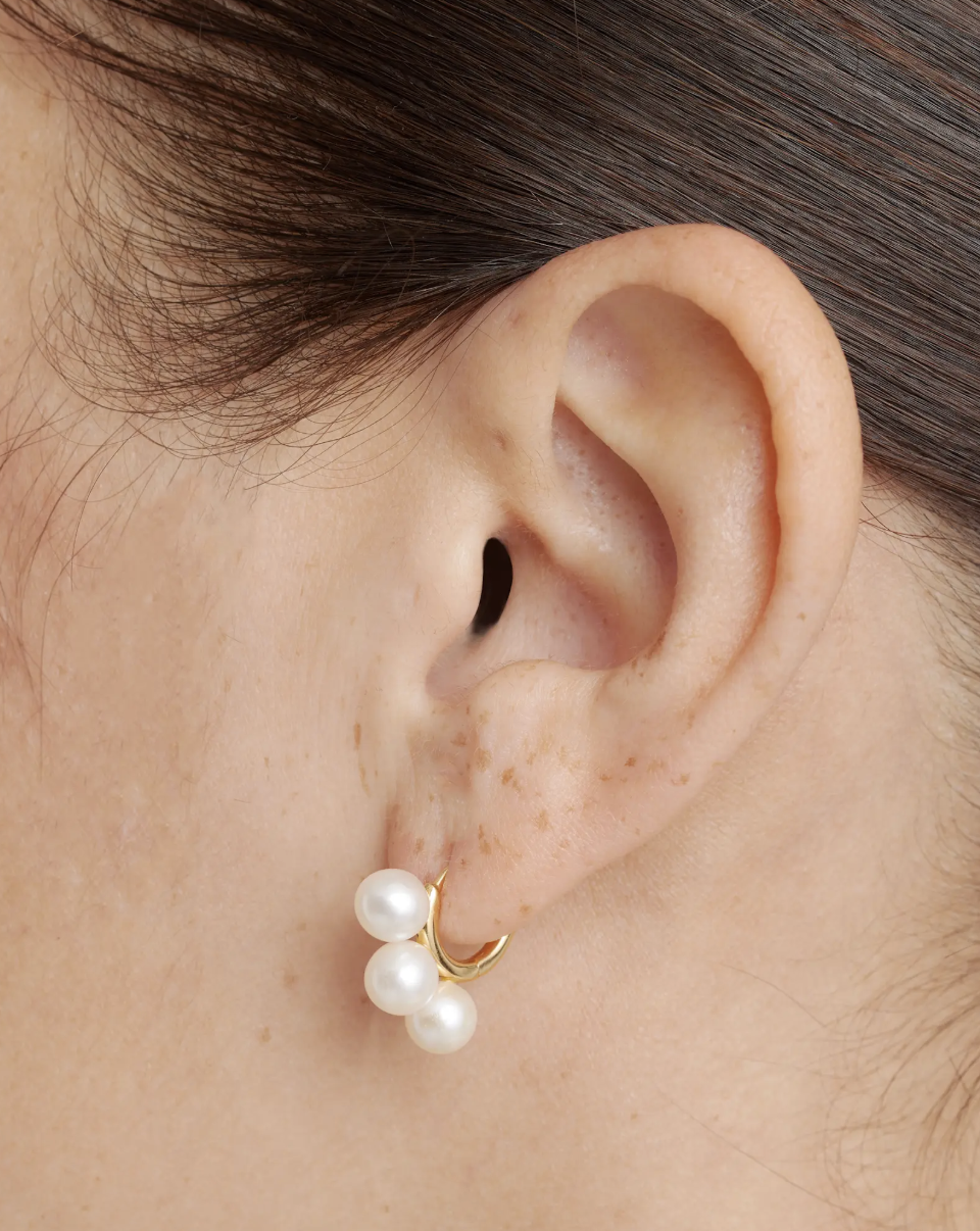 Close-up of a person’s ear wearing a gold hoop earring with three white pearls attached.