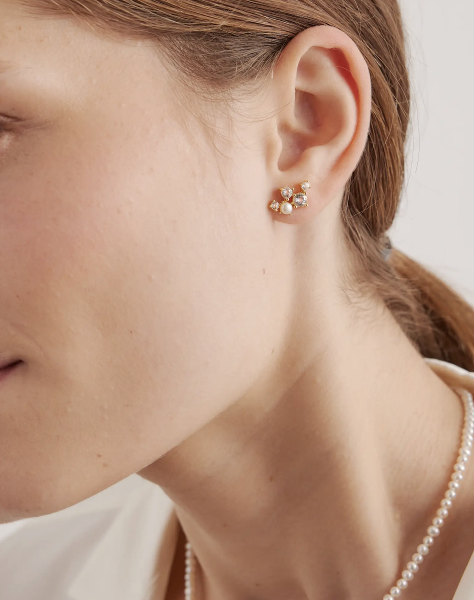 Close-up of a person's ear wearing a gold earring with multiple small gemstones. The person has light skin and brown hair, and is also wearing a pearl necklace.