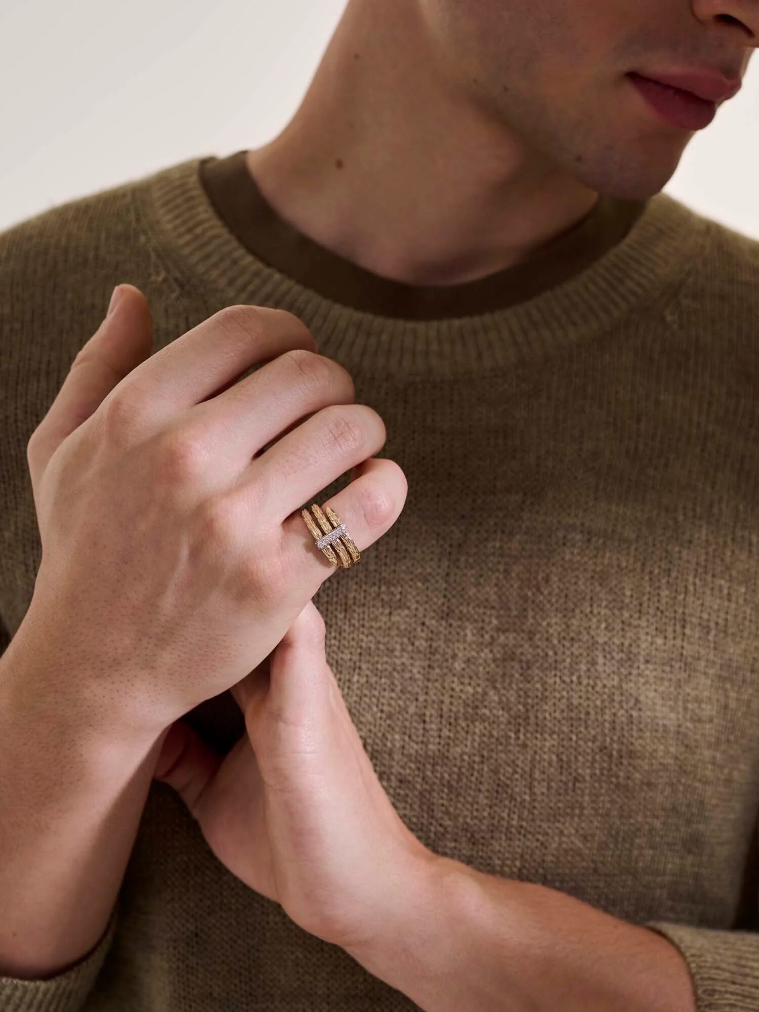 A person wearing a brown knit sweater and a gold ring on their finger, with hands held close to their chest against a plain background.