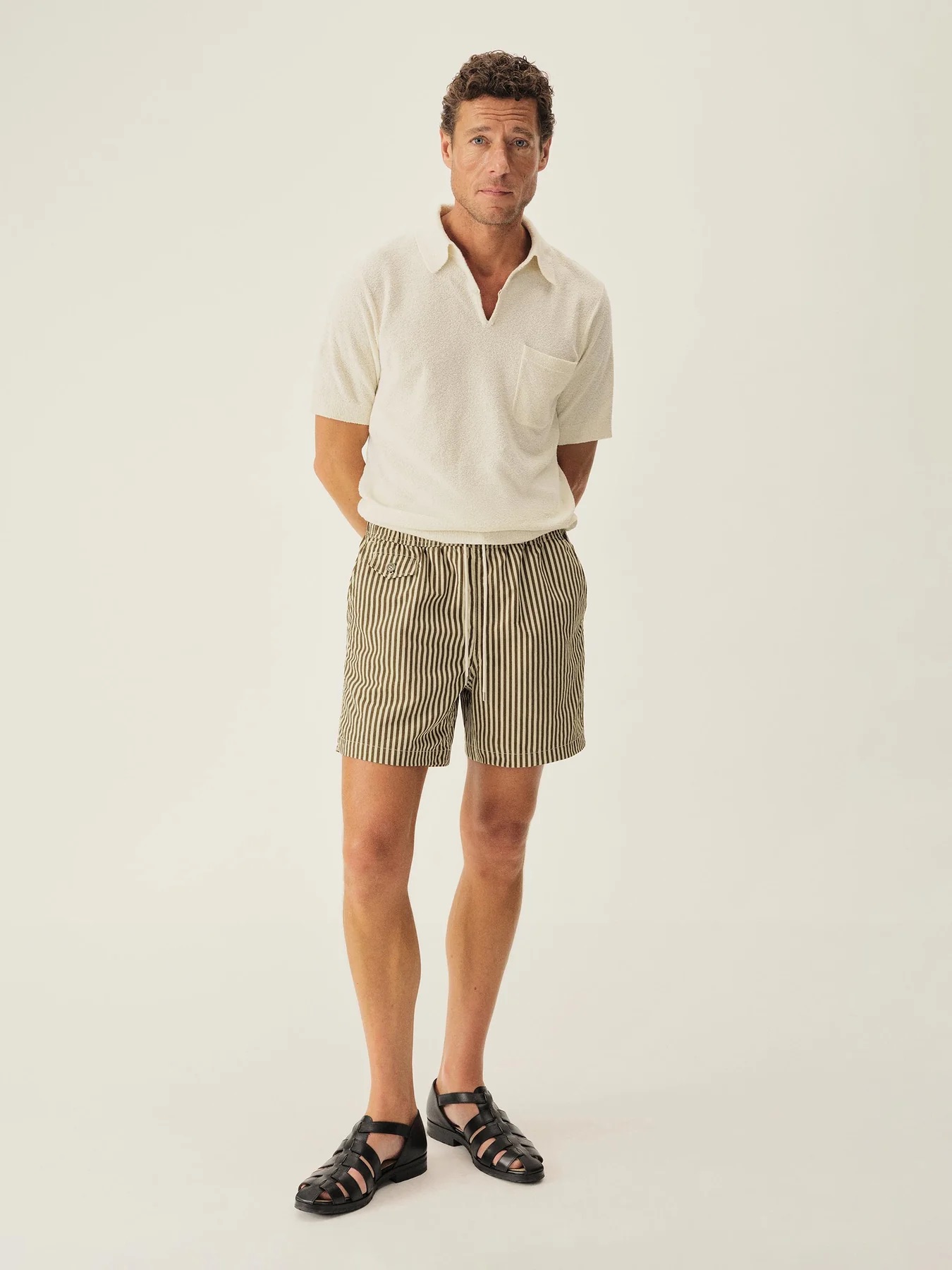 A man stands against a plain background wearing a short-sleeve white shirt, striped shorts, and black sandals with his hands behind his back.