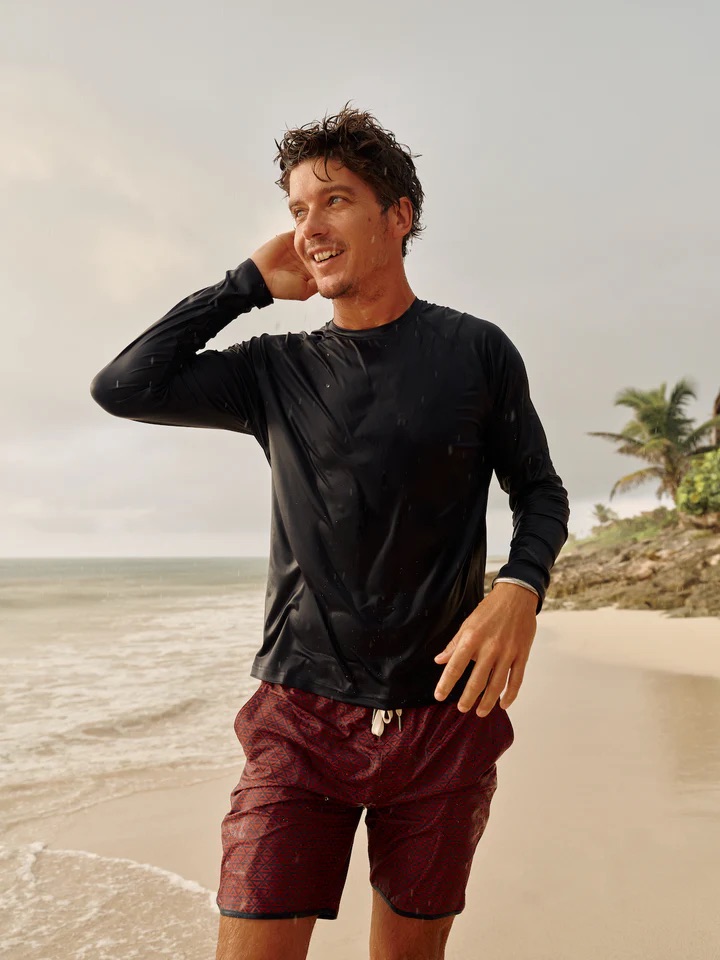 Man wearing a black long-sleeve shirt and red swim trunks stands on a sandy beach near the water, with palm trees and ocean in the background.