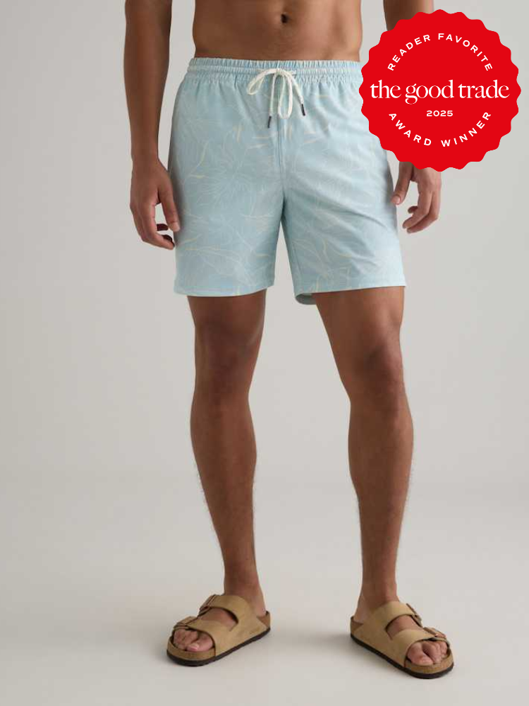 A person stands wearing light blue swim trunks with a subtle pattern and tan sandals. A red badge in the corner reads "The Good Trade Reader Favorite Award Winner 2025.