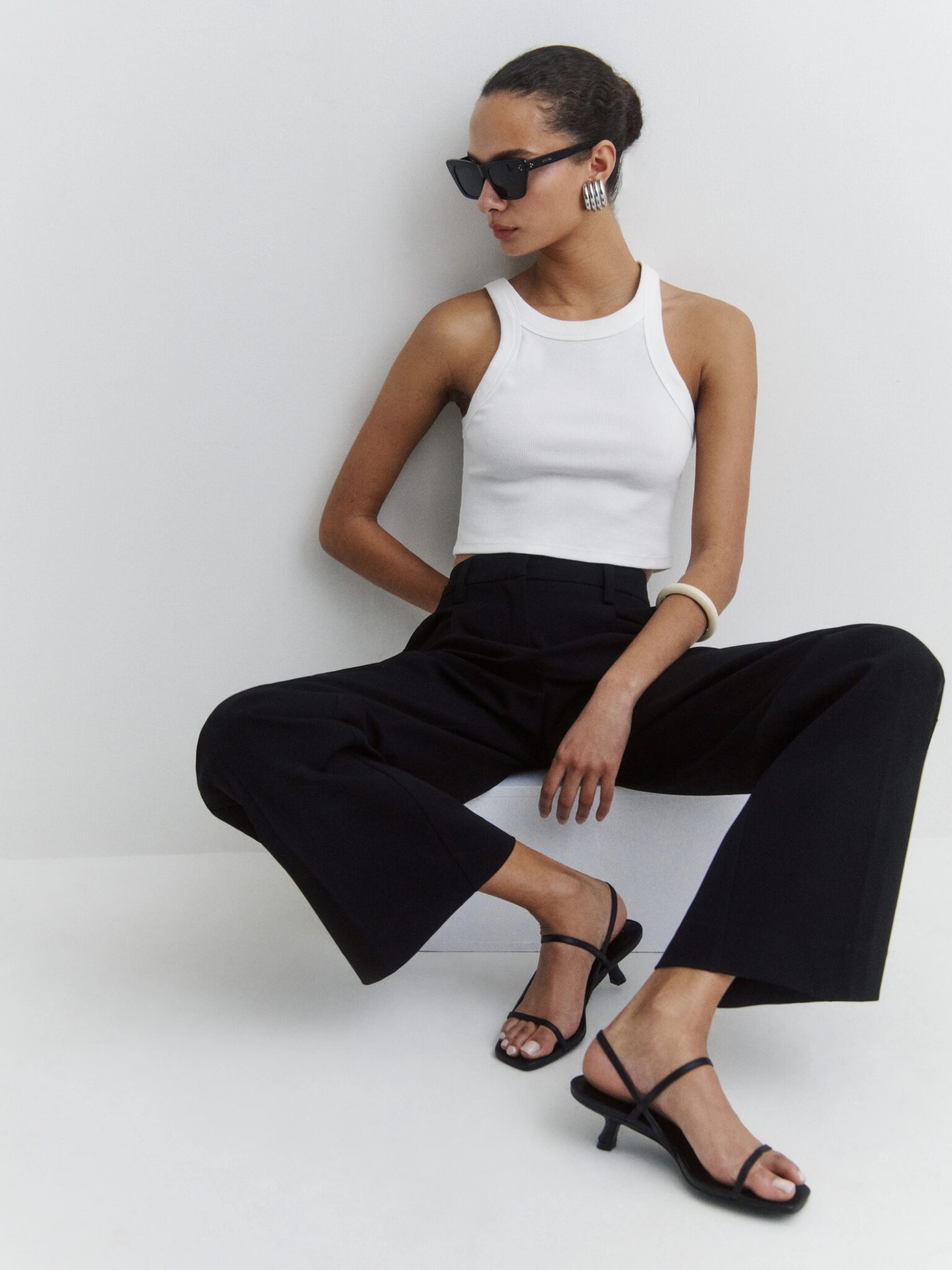 Woman wearing a white sleeveless top, black wide-leg pants, black heeled sandals, sunglasses, and statement earrings, seated against a plain white background.