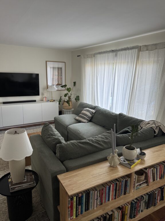 A living room with a sectional sofa, a TV mounted on the wall, a bookshelf filled with books, indoor plants, and sheer curtains covering large windows.