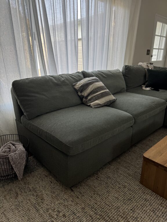 A dark gray sectional sofa with two cushions and a striped pillow sits in front of a window with sheer curtains, next to a wooden table and a basket with a blanket.