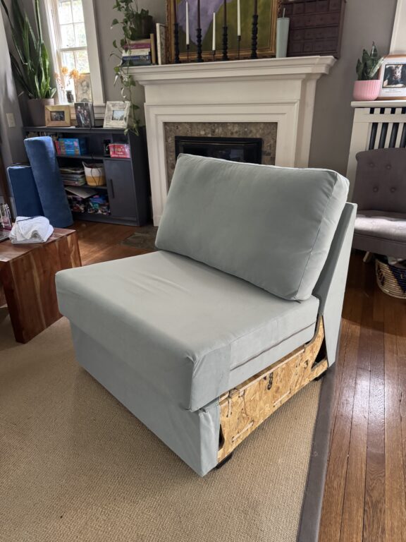A light gray cushioned chair with exposed wooden frame sits in a living room with hardwood floors, a fireplace, and various furniture and decor items.