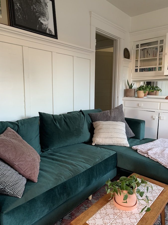 A green velvet sectional sofa with assorted pillows and a knitted throw, a wooden coffee table with a potted plant, and white built-in cabinets in a cozy living room.