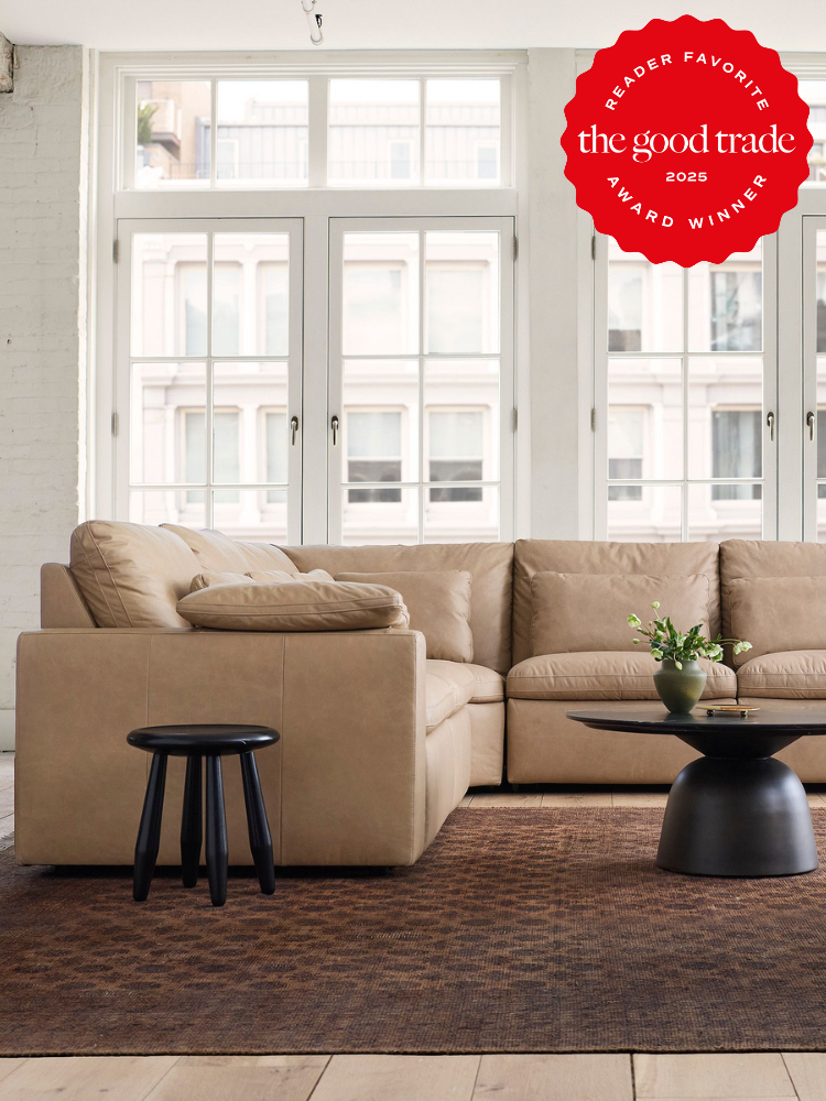 A beige sectional sofa sits on a brown rug in a bright living room with large windows; a small black stool and a round coffee table are nearby. “The Good Trade 2025 Award Winner” badge is displayed.