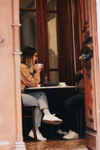 A woman sits at a small round table in a wooden café, holding a mug and talking to someone whose face is not visible.