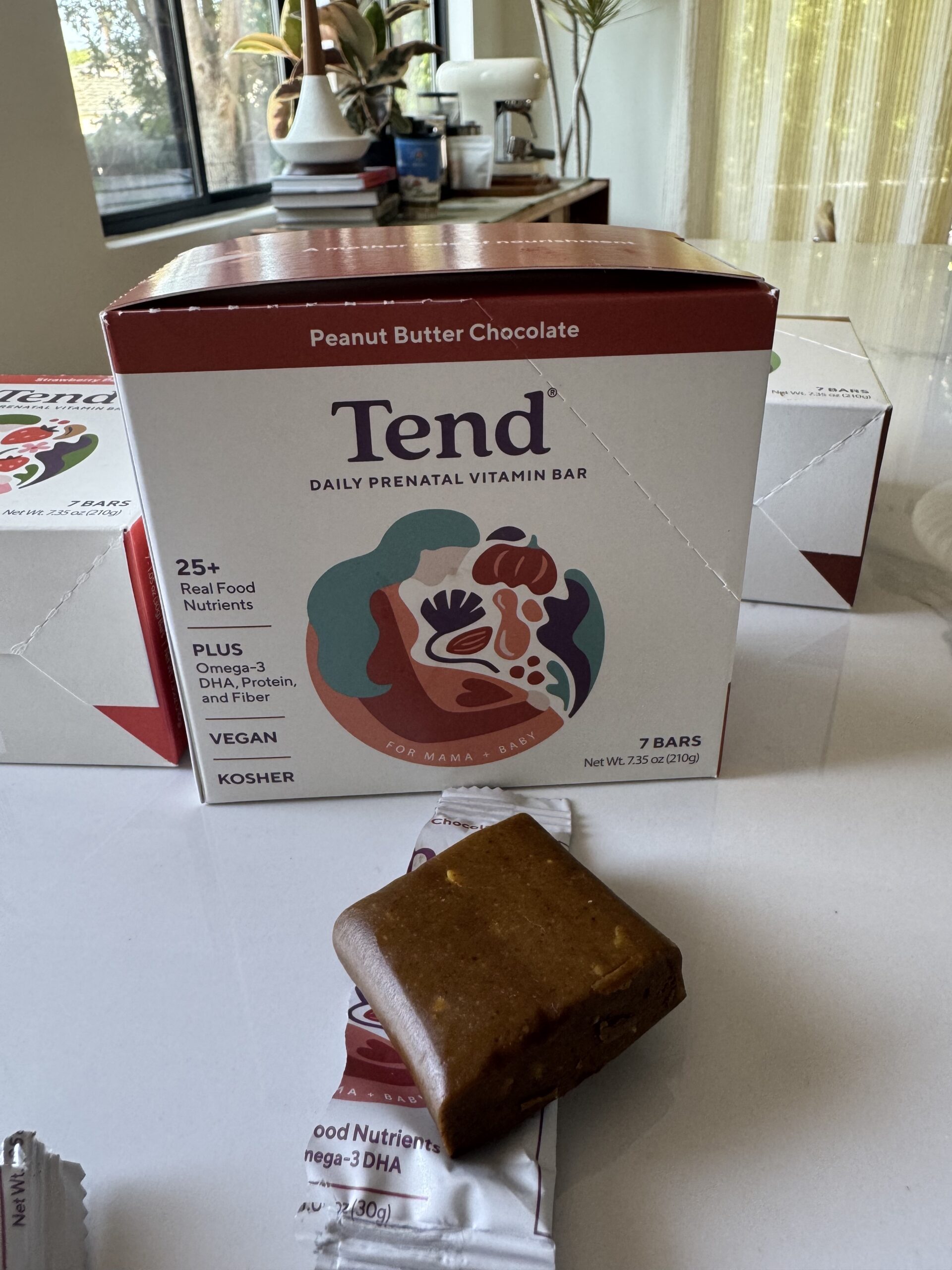 A box of Tend peanut butter chocolate prenatal vitamin bars sits on a table, with one unwrapped bar placed in front of the box.