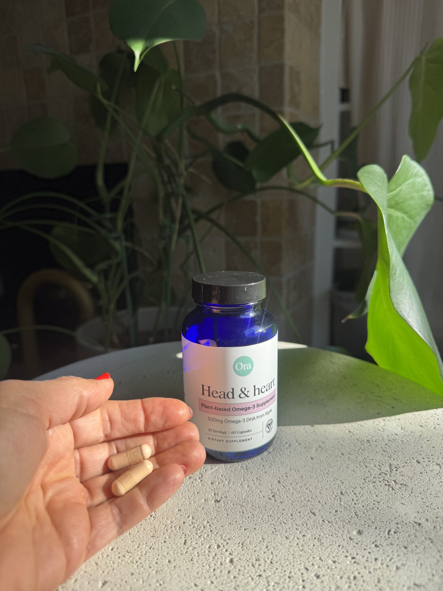 A hand holds three capsules next to a blue bottle labeled "Head & Heart Plant-Based Omega 3 Supplement" on a sunlit surface with green plants in the background.