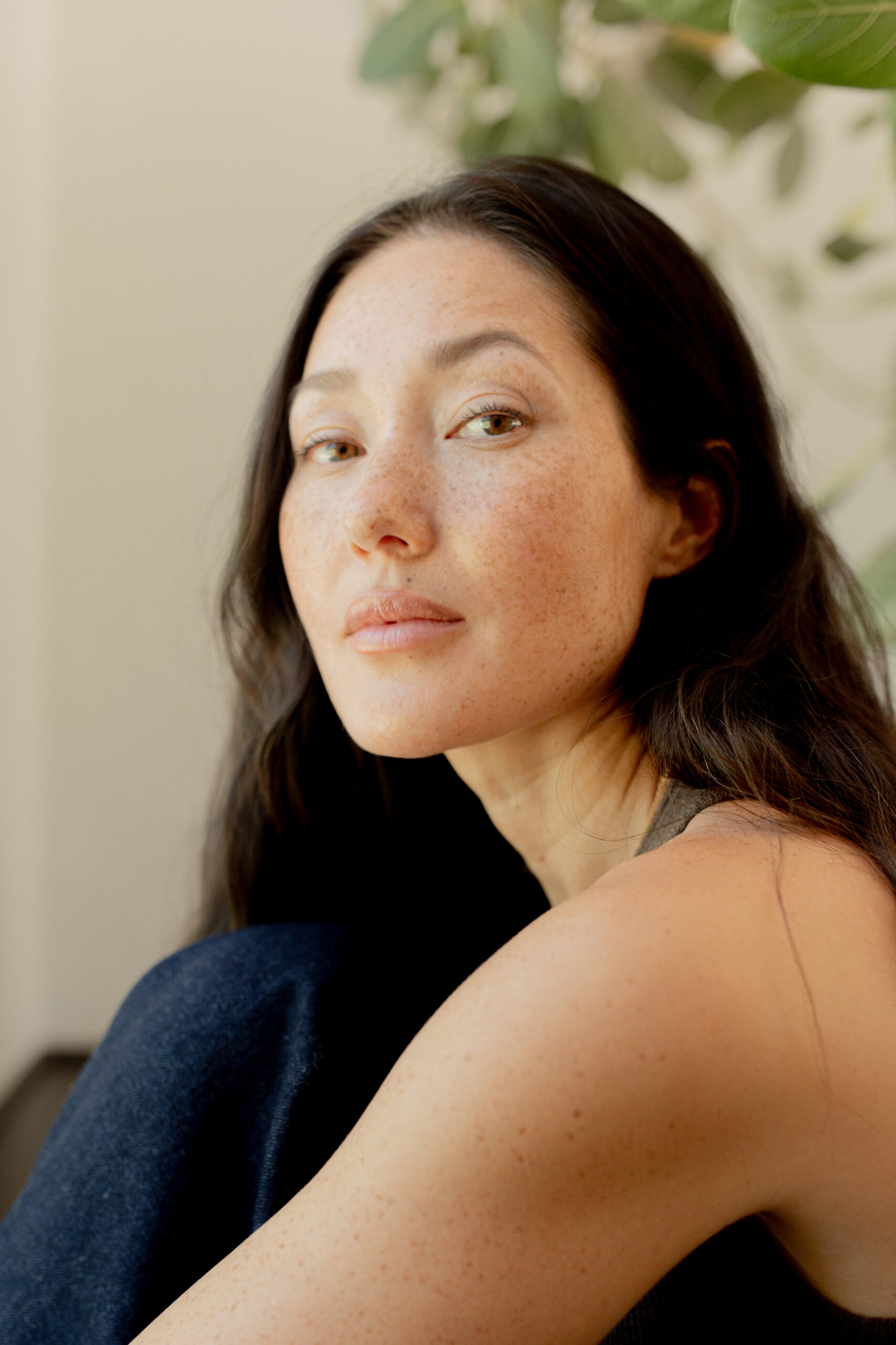 A woman with long dark hair and freckles looks at the camera, sitting indoors with a plant in the background.