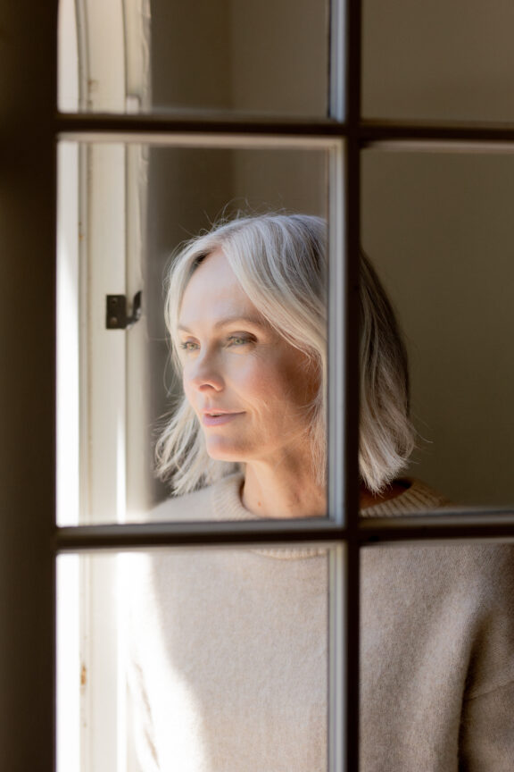 A woman with gray hair and a beige sweater looks out a sunlit window, framed by glass panes.