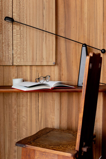 A wooden desk with an open book, eyeglasses, a cup, and a modern black desk lamp, set against a wood-paneled wall and accompanied by a wooden chair.