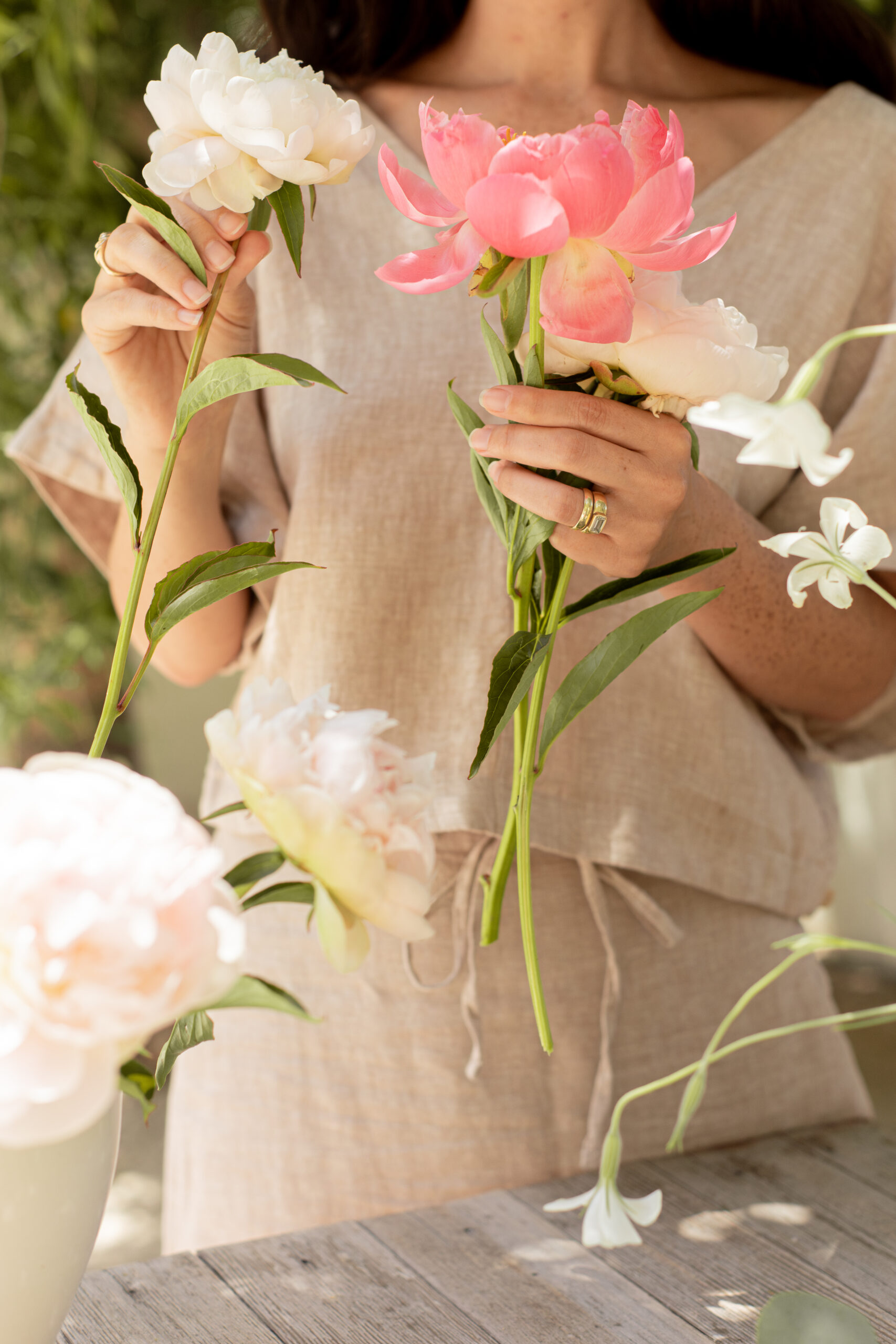 A woman in a beige dress holds pink and white flowers while standing by a wooden table with more flowers in a vase.