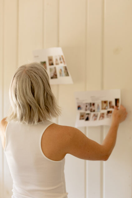 A person with gray hair in a white sleeveless top holds and looks at two photo sheets taped to a white paneled wall.