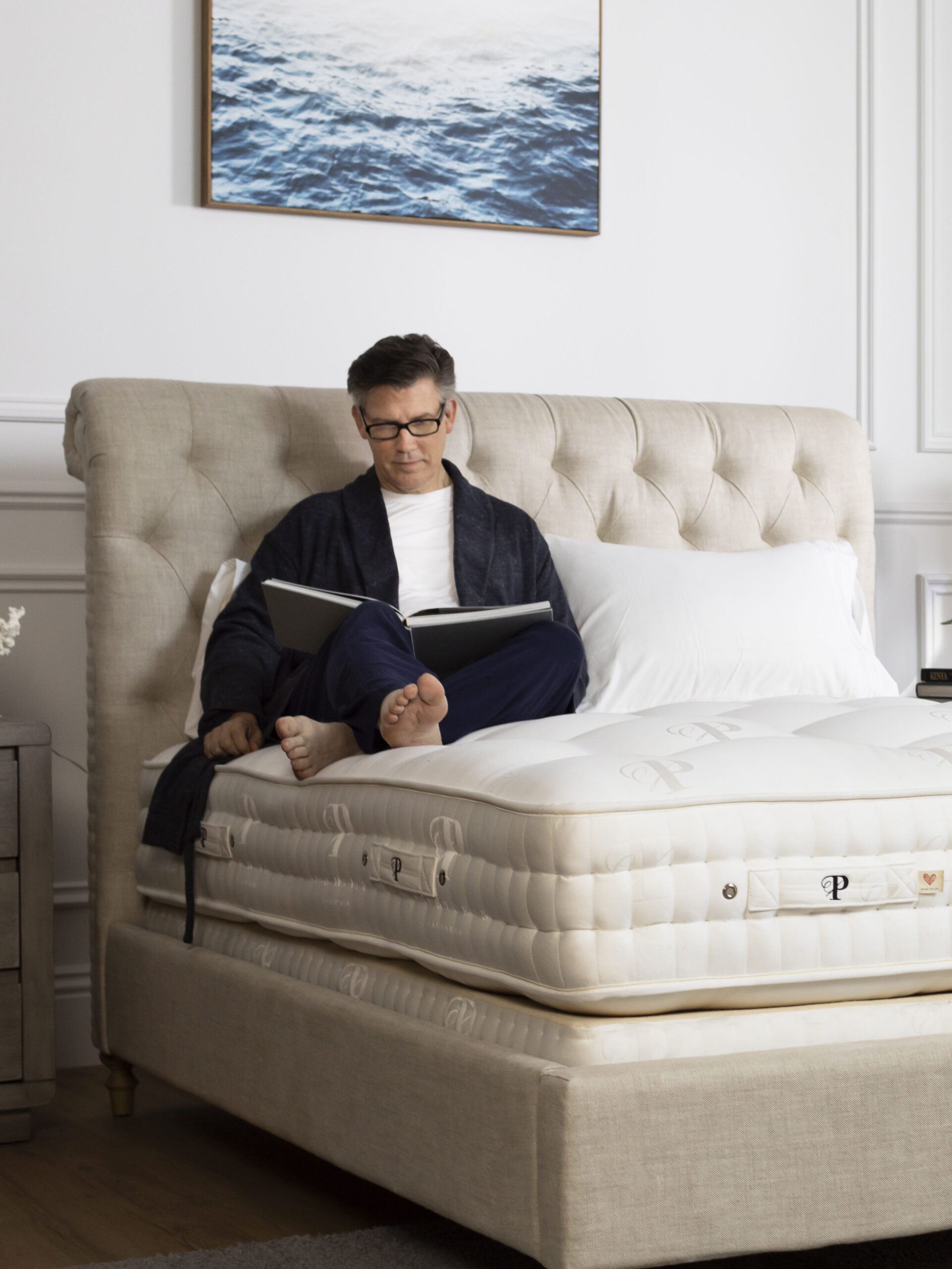 A person in glasses sits on a bed reading a book in a bright, neatly decorated bedroom with a large window and natural light.