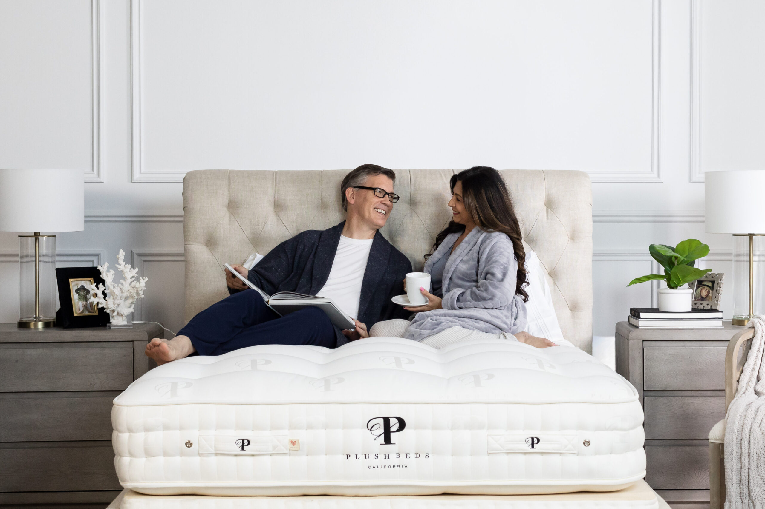 A man and woman sit on a bed in a modern bedroom, wearing robes, reading and drinking from mugs, with two nightstands and lamps on either side.