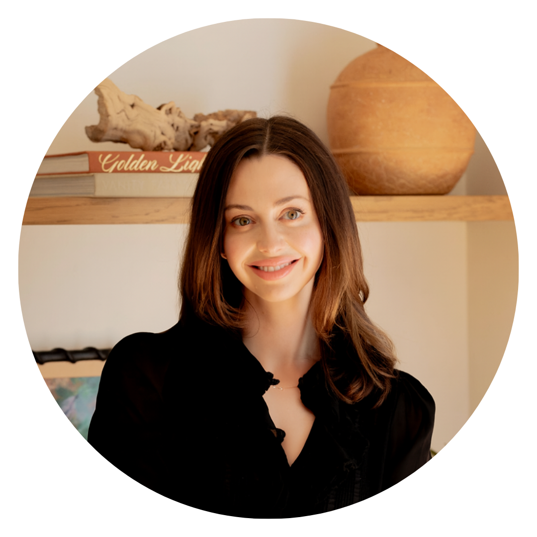 A woman with long brown hair and a black top stands in front of a shelf with books, a ceramic pot, and a piece of driftwood, smiling at the camera.
