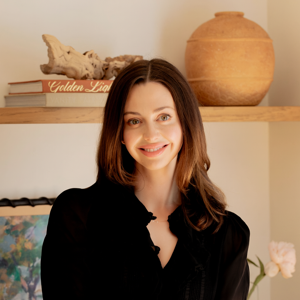 A woman with long brown hair and a black top smiles in front of a wooden shelf with books, a clay pot, driftwood, and a framed picture.