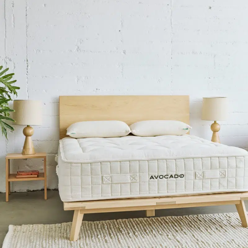 A minimalist bedroom with a wooden bed frame, a white Avocado mattress, two pillows, matching bedside tables, lamps, a plant, and a white textured rug on a concrete floor.