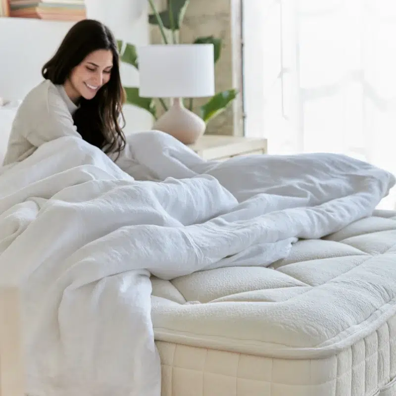 A woman with long dark hair sits up in bed, smiling, under a white comforter in a bright, modern bedroom.