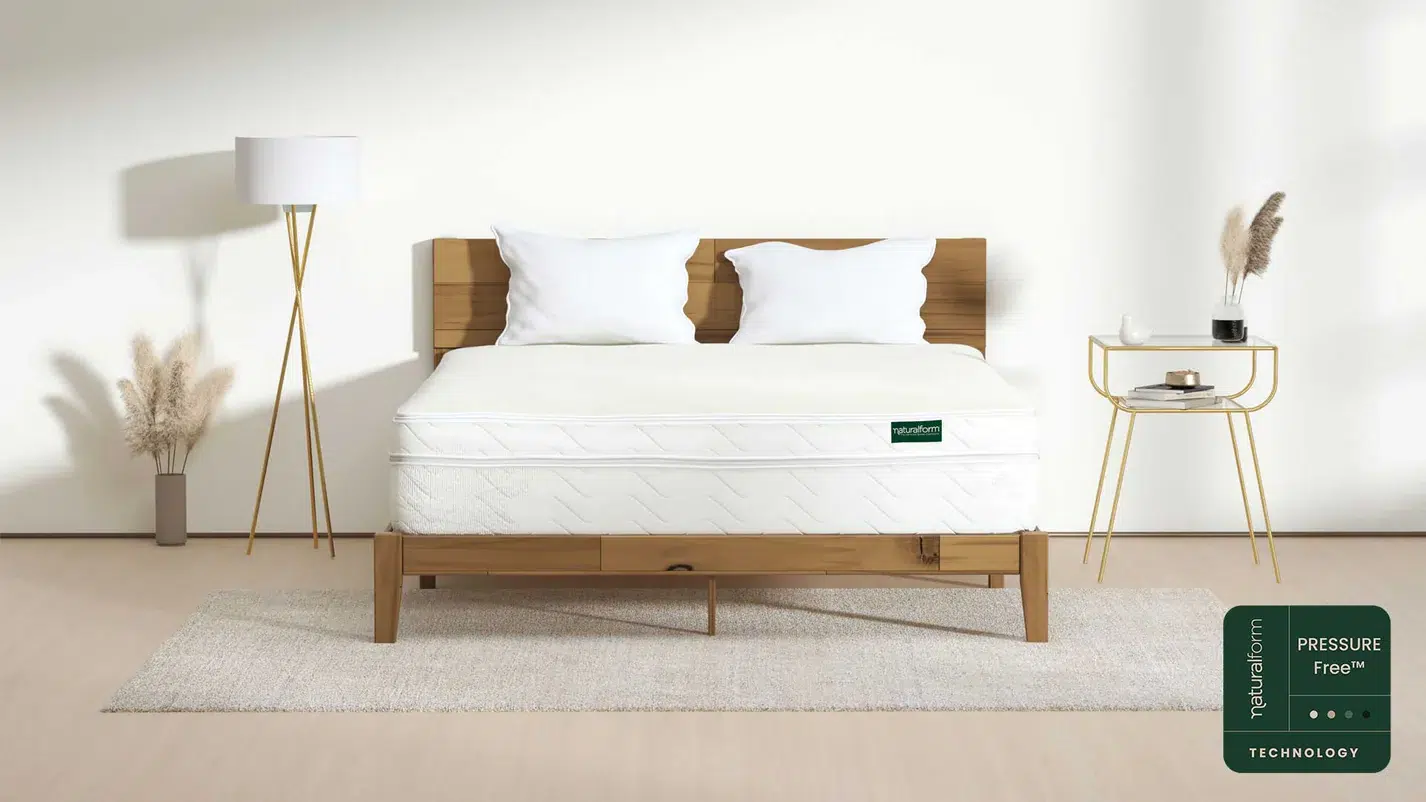 A neatly made bed with white bedding and pillows on a wooden frame, flanked by a floor lamp and a side table, in a minimalist bedroom. Naturafirm Pressure Free Technology logo in corner.