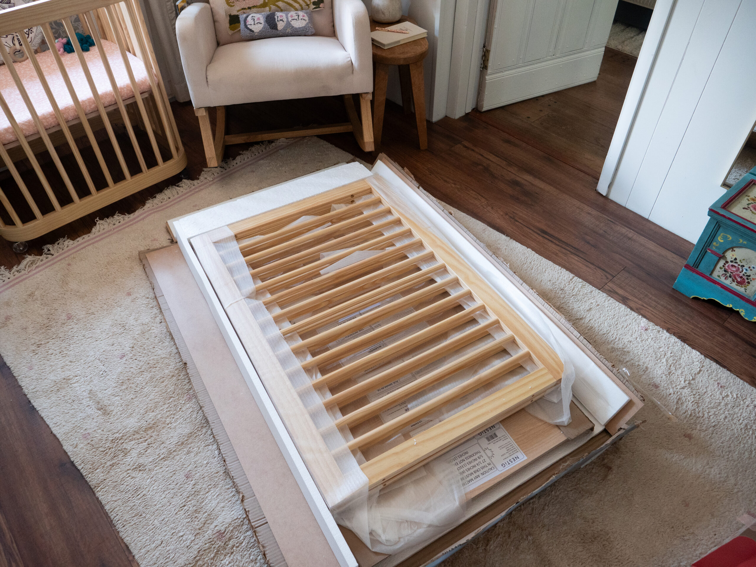 A wooden crib side panel lies on packaging materials and carpet, next to a crib, armchair, and stool in a nursery room.
