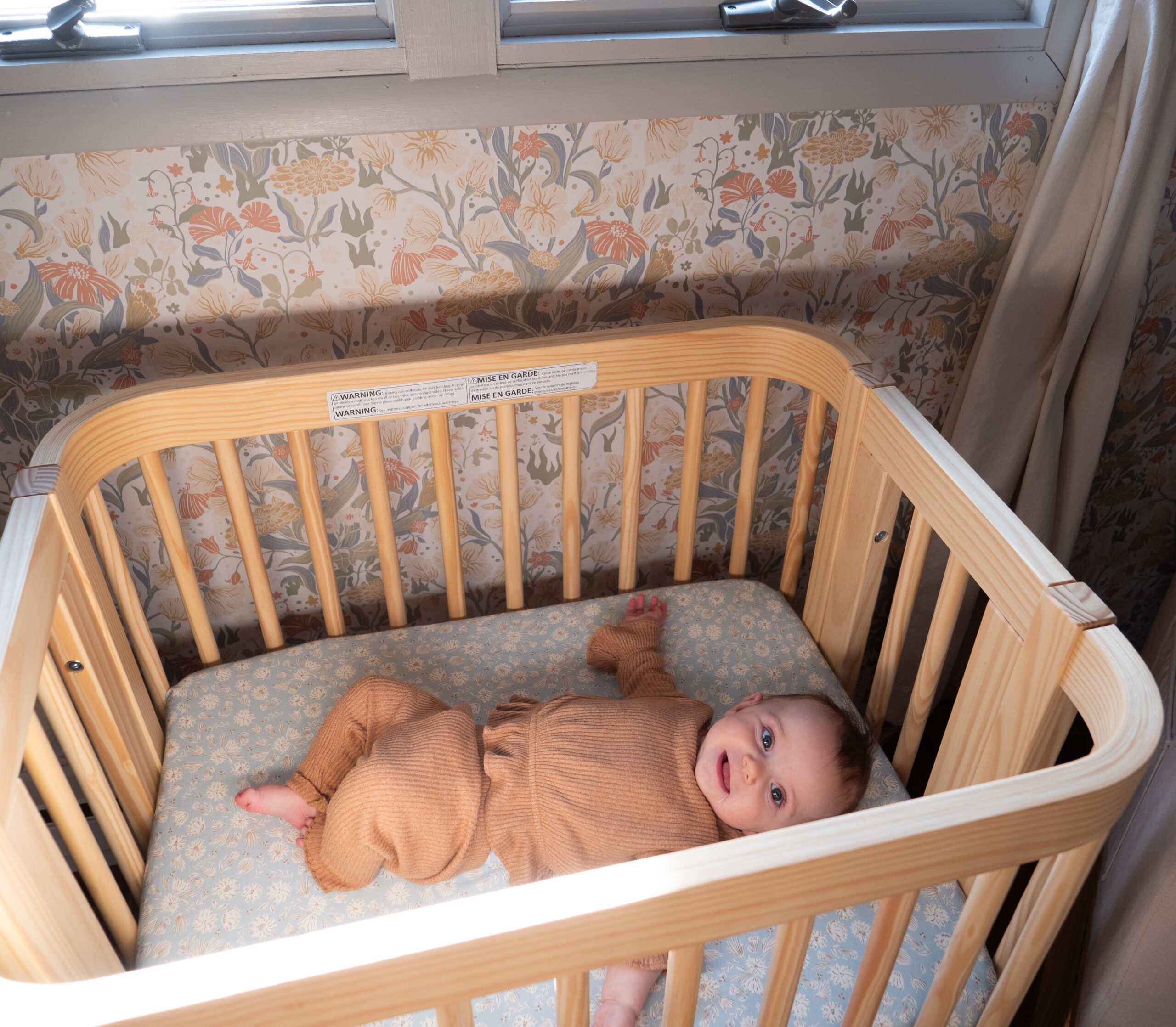 A baby in a tan outfit lies on their back in a wooden crib with floral wallpaper and a window in the background.
