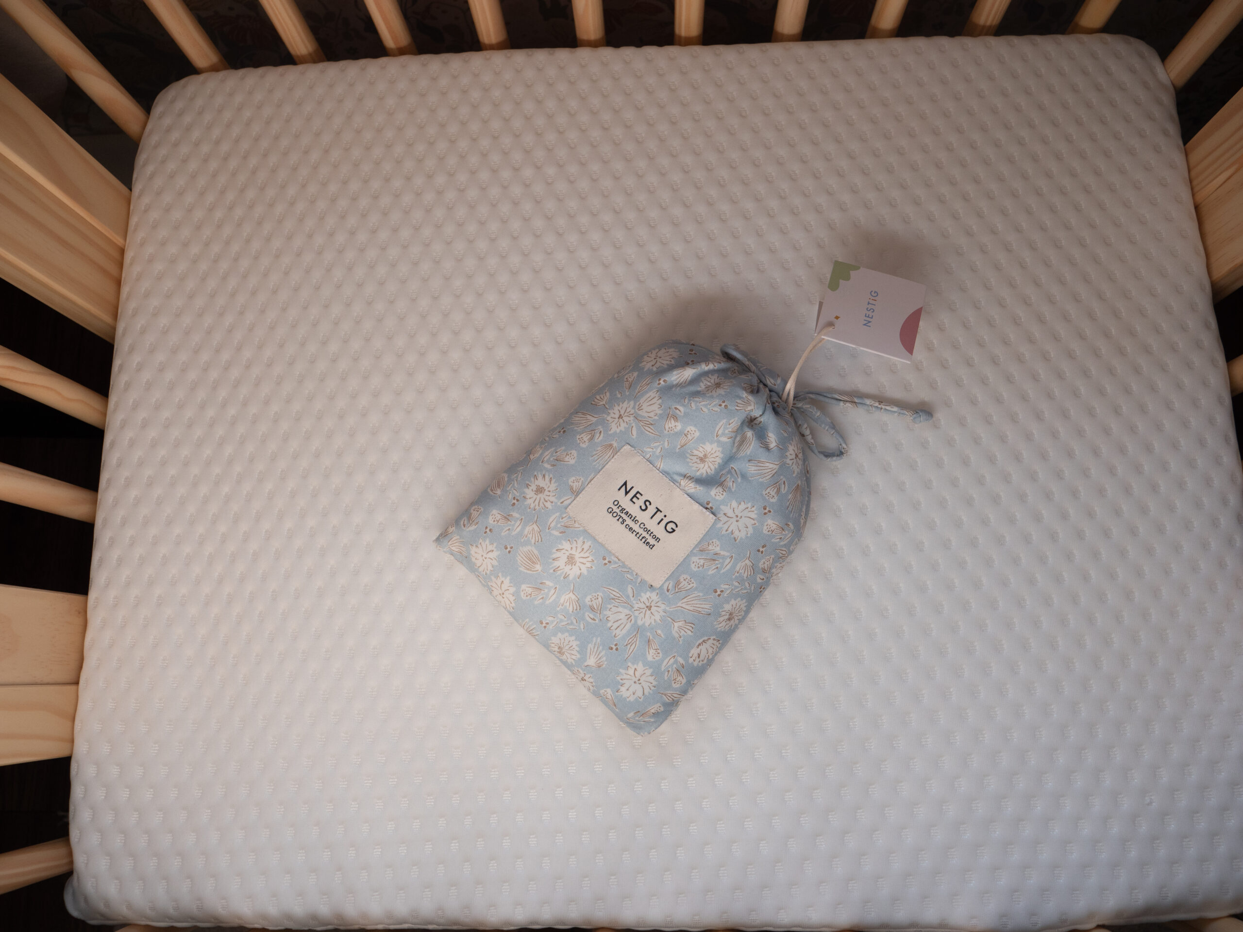 A small blue drawstring bag labeled "NESTIG" rests on a white fitted crib mattress inside a wooden crib.