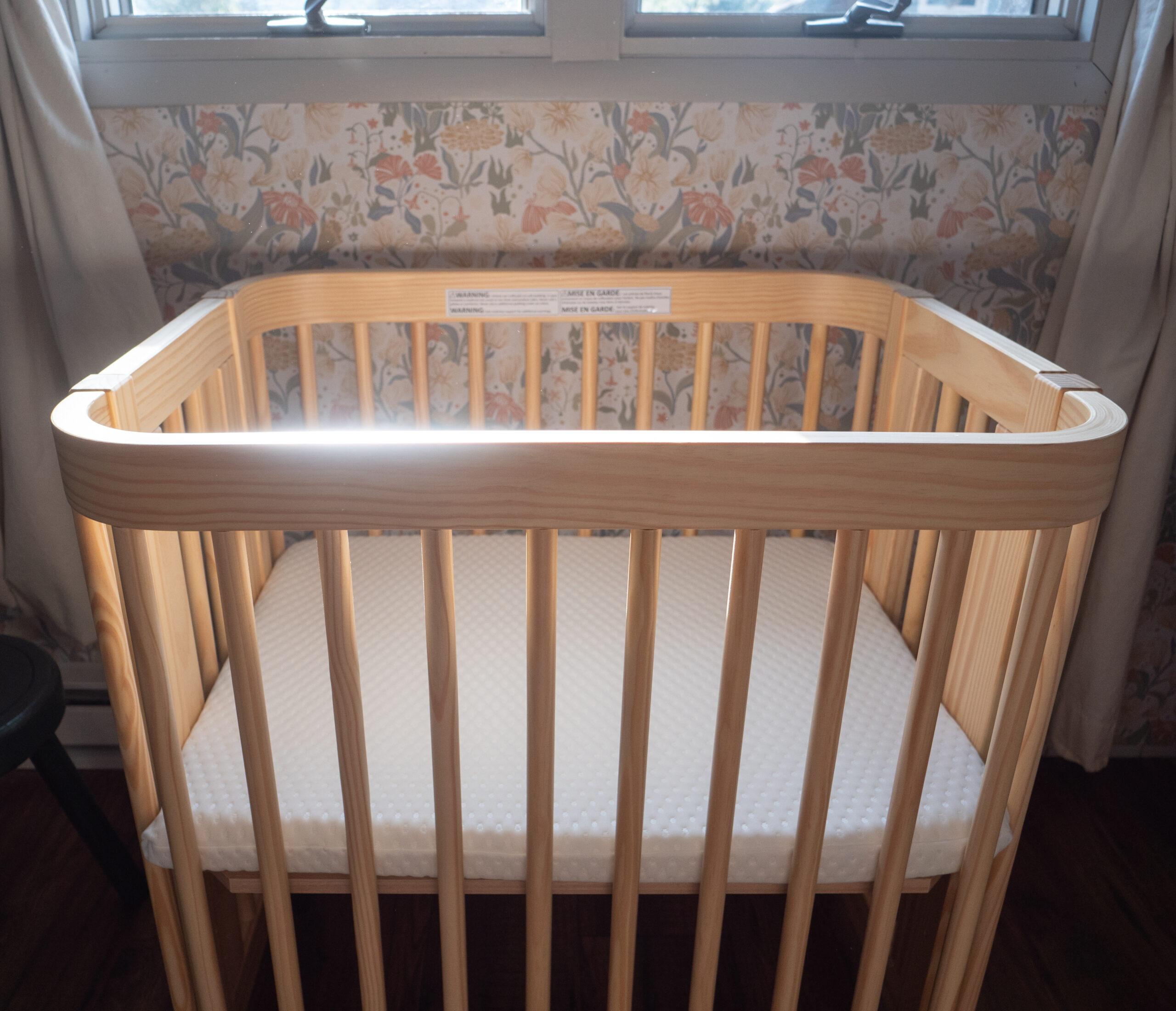 A wooden baby crib with a white mattress is placed beneath a window in a room with floral wallpaper.