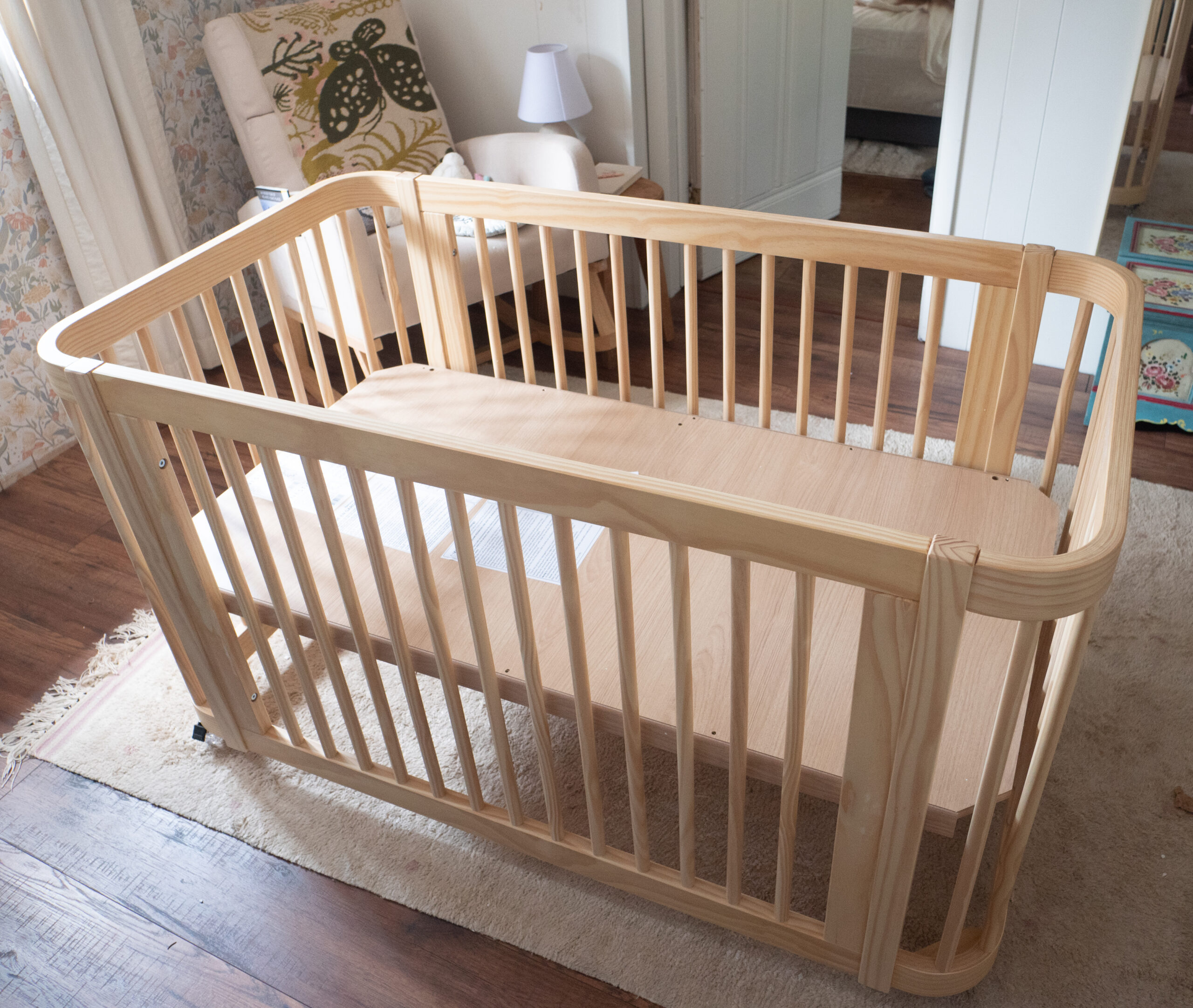 A wooden baby crib with a flat base stands in a cozy, well-lit room with wooden floors, a rug, and a chair nearby.