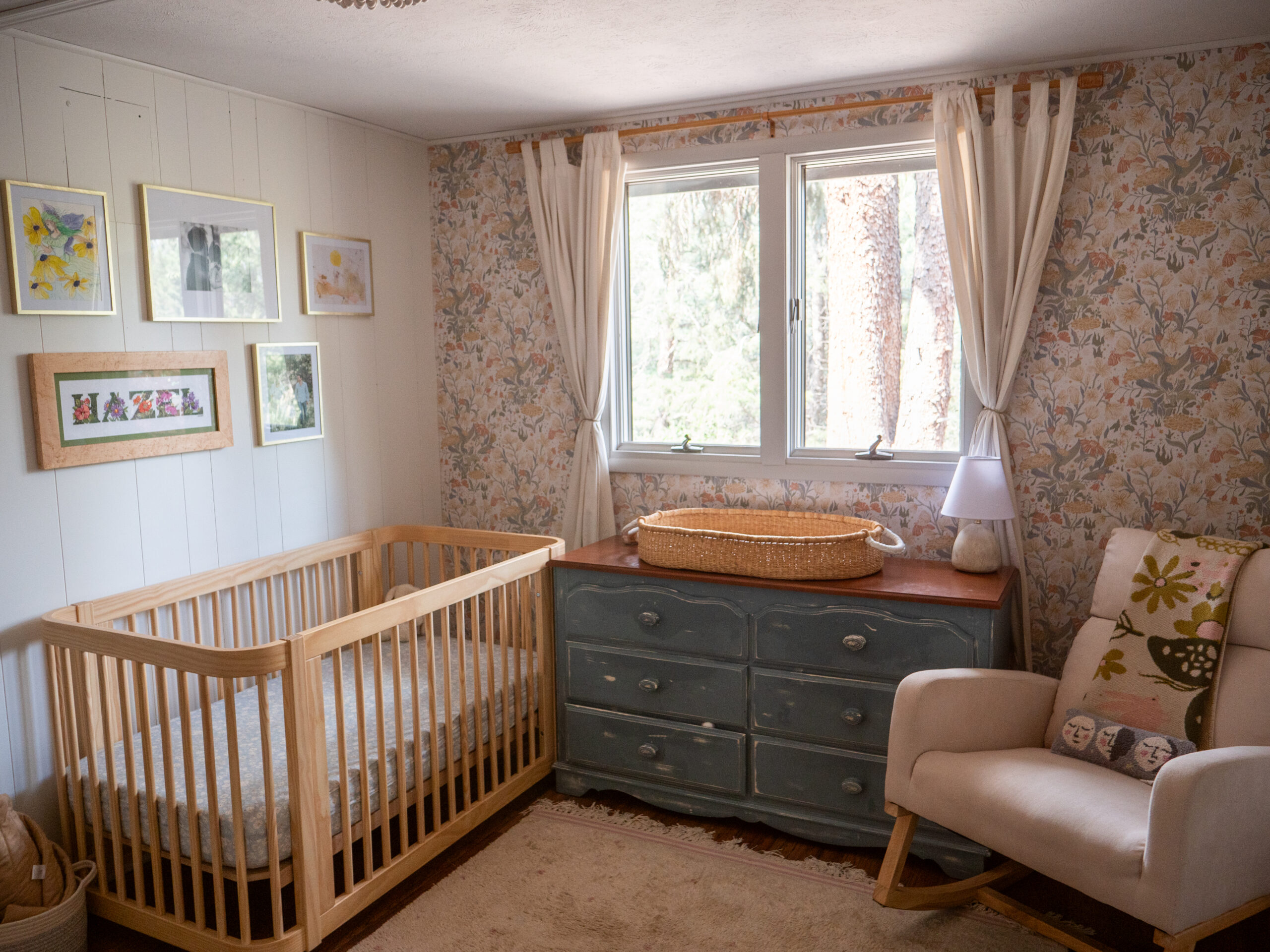A cozy nursery with a wooden crib, changing table with basket, armchair, framed art on white walls, and floral wallpaper, all softly lit by a window with cream curtains.