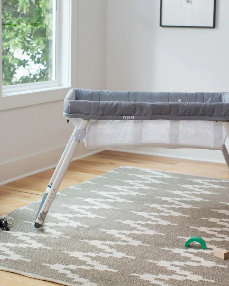 A gray and white baby bassinet stands on a patterned rug in a bright room with wooden floors and a large window. Toys are scattered on the rug.