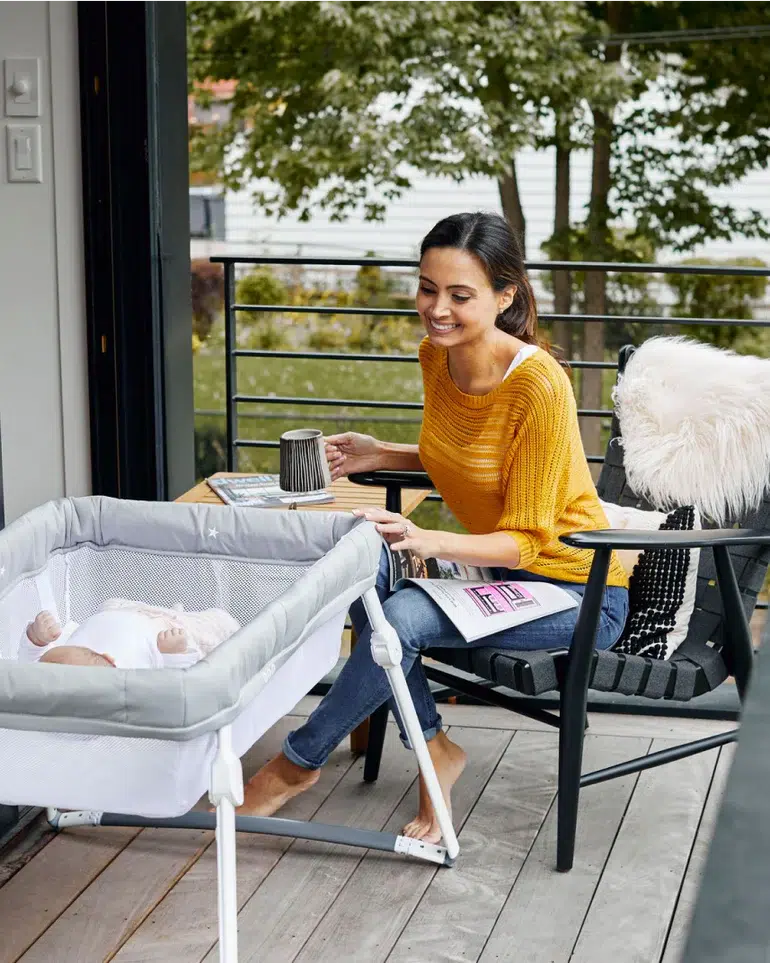 A woman sits on a porch holding a mug and magazine, smiling at a baby lying in a bassinet beside her. Trees and a railing are visible in the background.