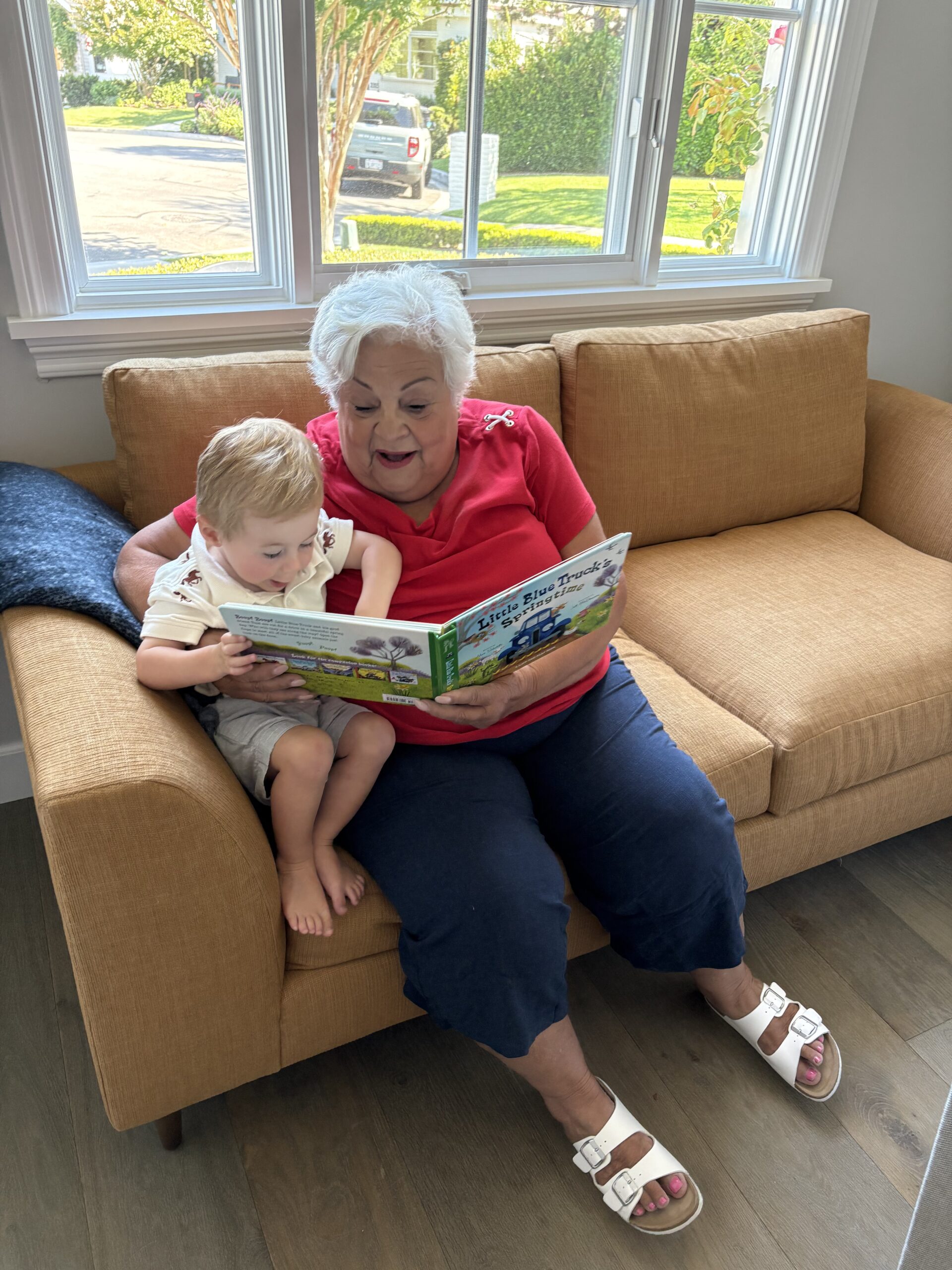 An older woman and a young child sit together on a tan couch, reading a children's book in a brightly lit room with large windows.
