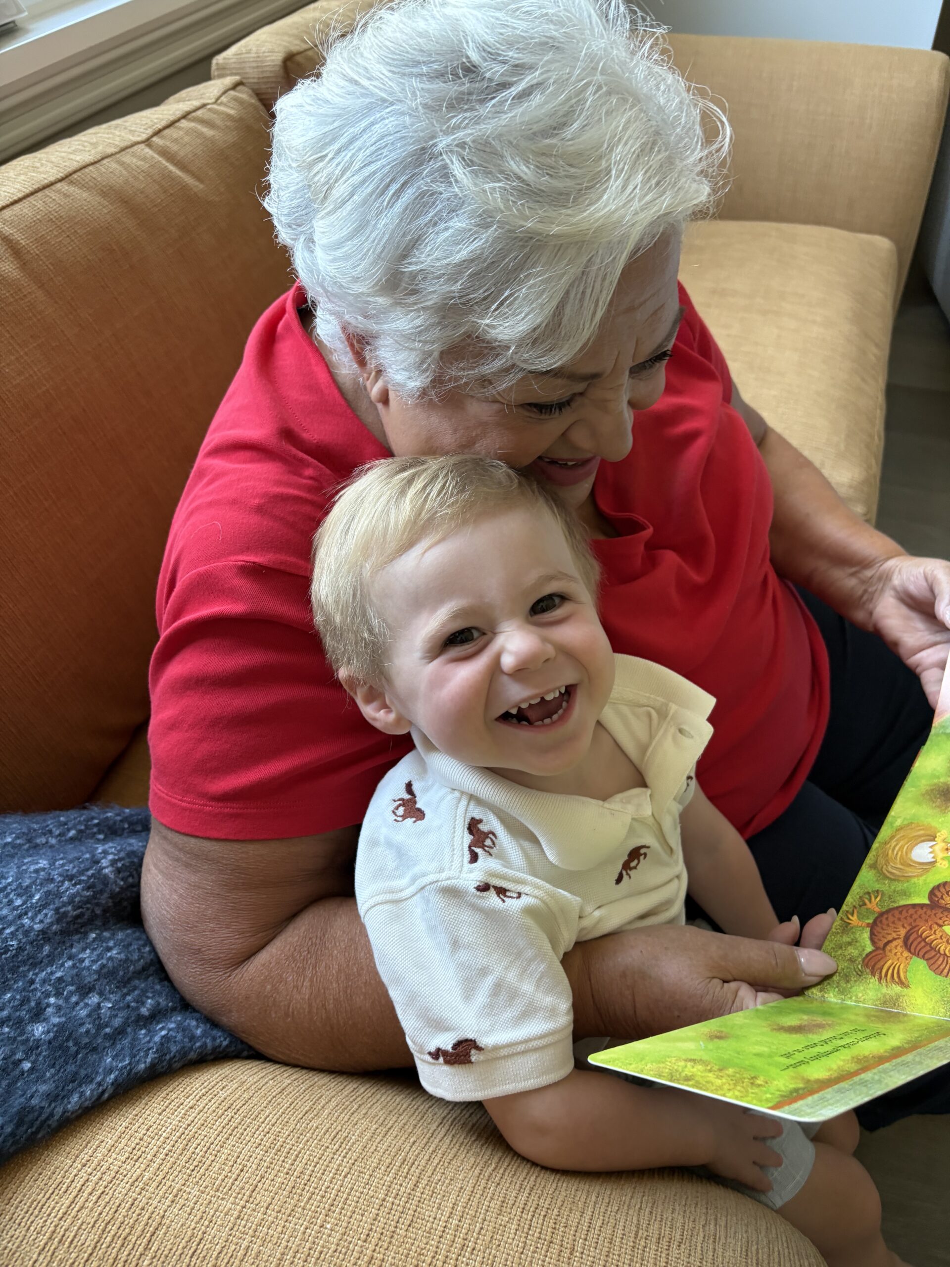 Older woman with short white hair and red shirt reads a picture book to a smiling toddler on her lap while sitting on a tan couch.