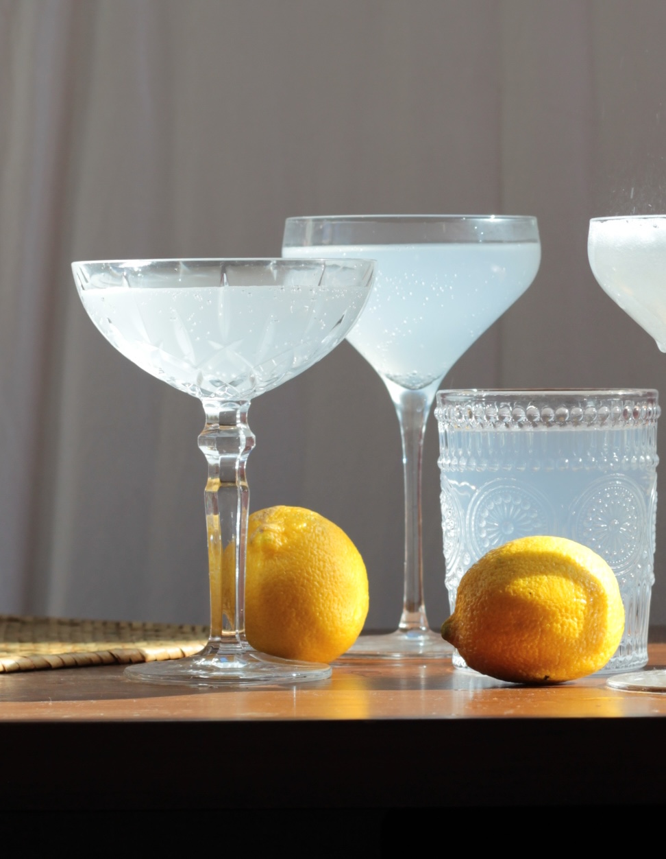 Four glasses with a clear, pale drink and two lemons are arranged on a sunlit wooden table, with a neutral-colored curtain in the background.