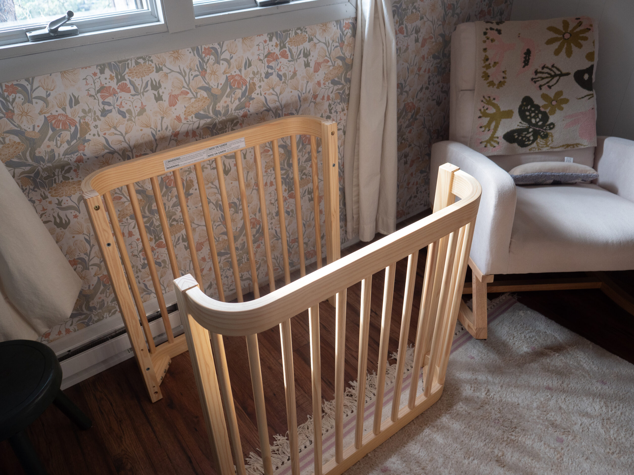 A partially assembled wooden crib frame sits on a rug in a nursery next to a white armchair and floral wallpaper.