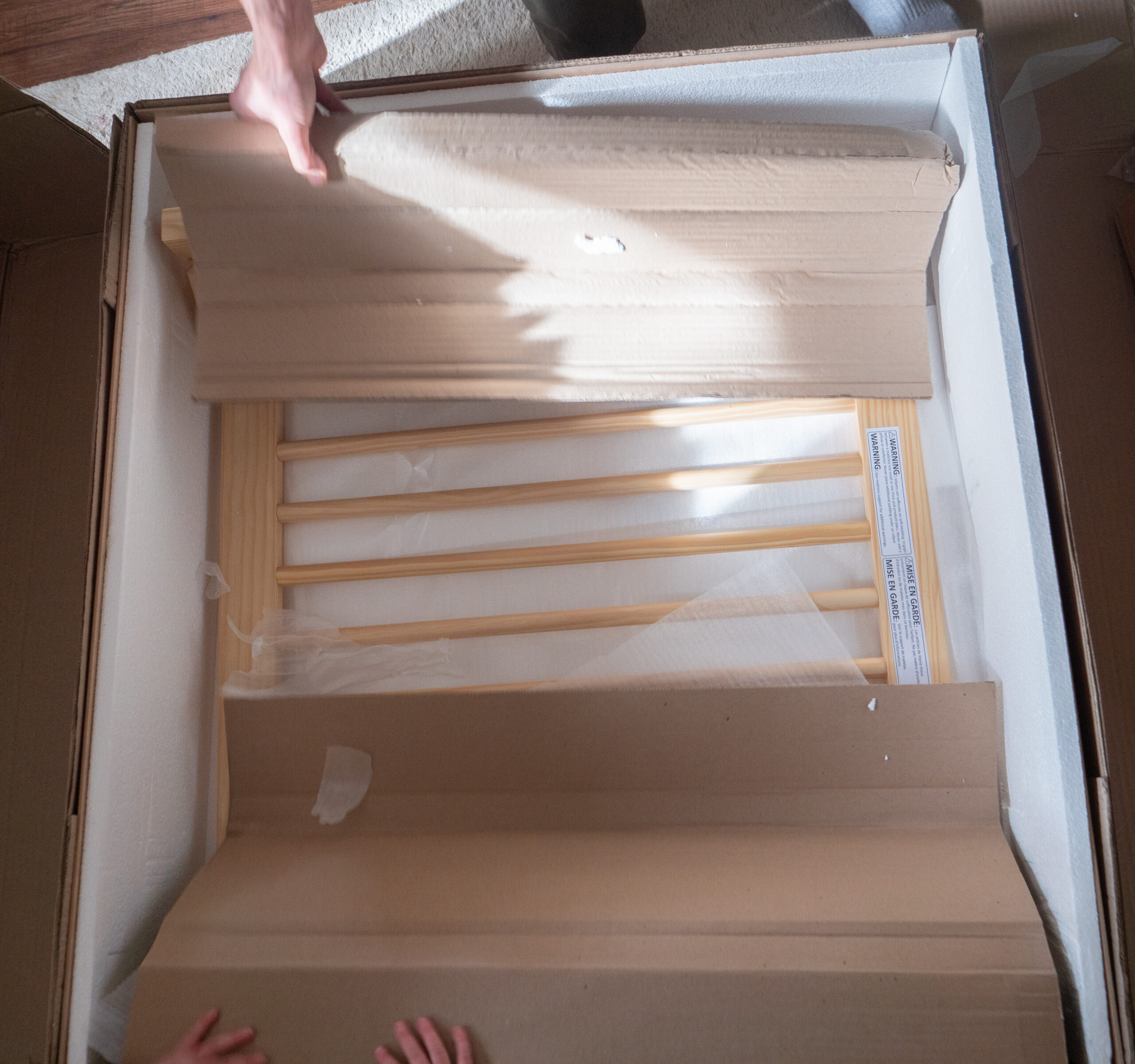 A person opens a cardboard box containing a wooden rack or crib side, partially covered by packaging material.