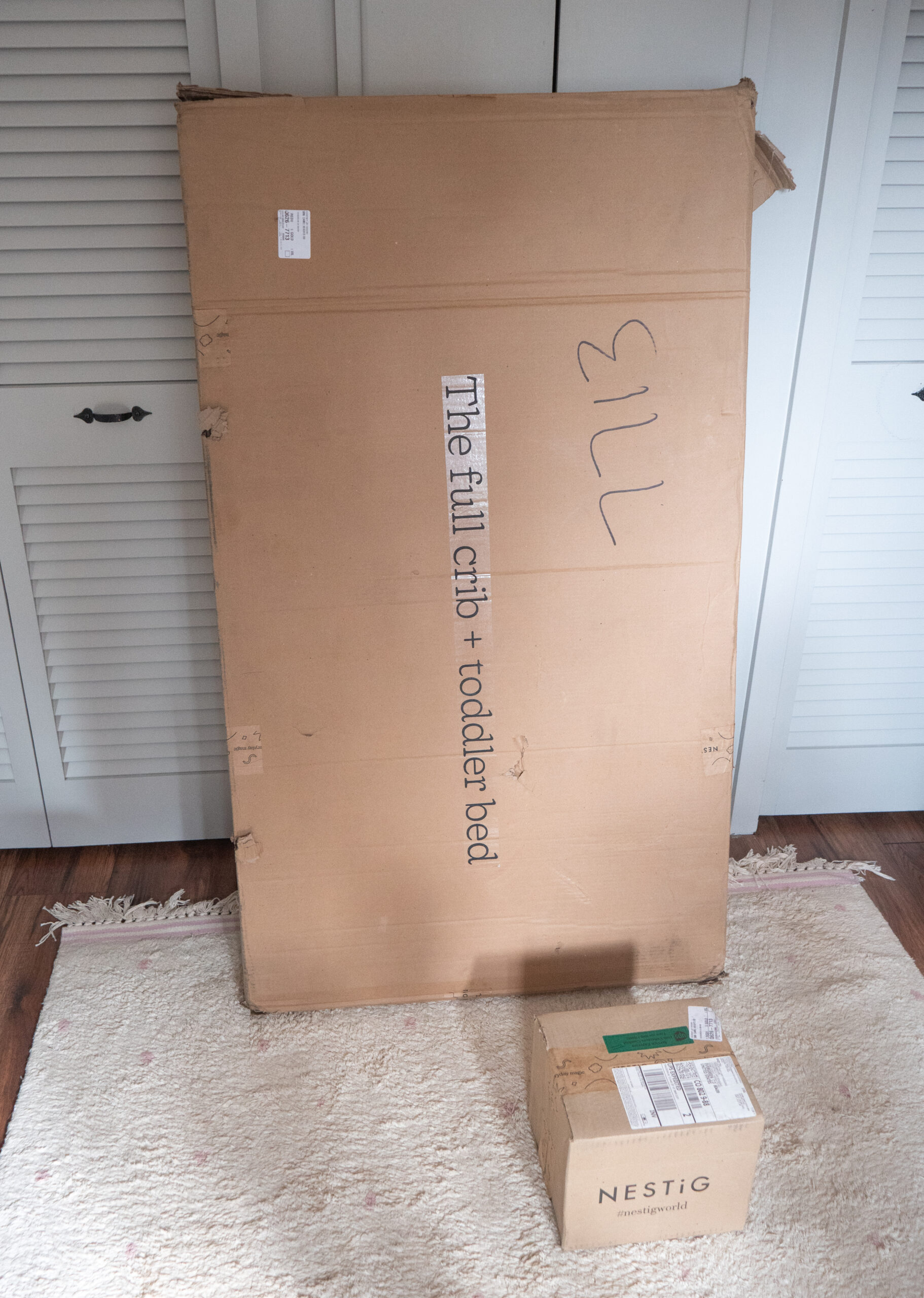 A large cardboard box labeled "The full crib + toddler bed" stands upright behind a smaller box labeled "NESTIG" on a light-colored rug.