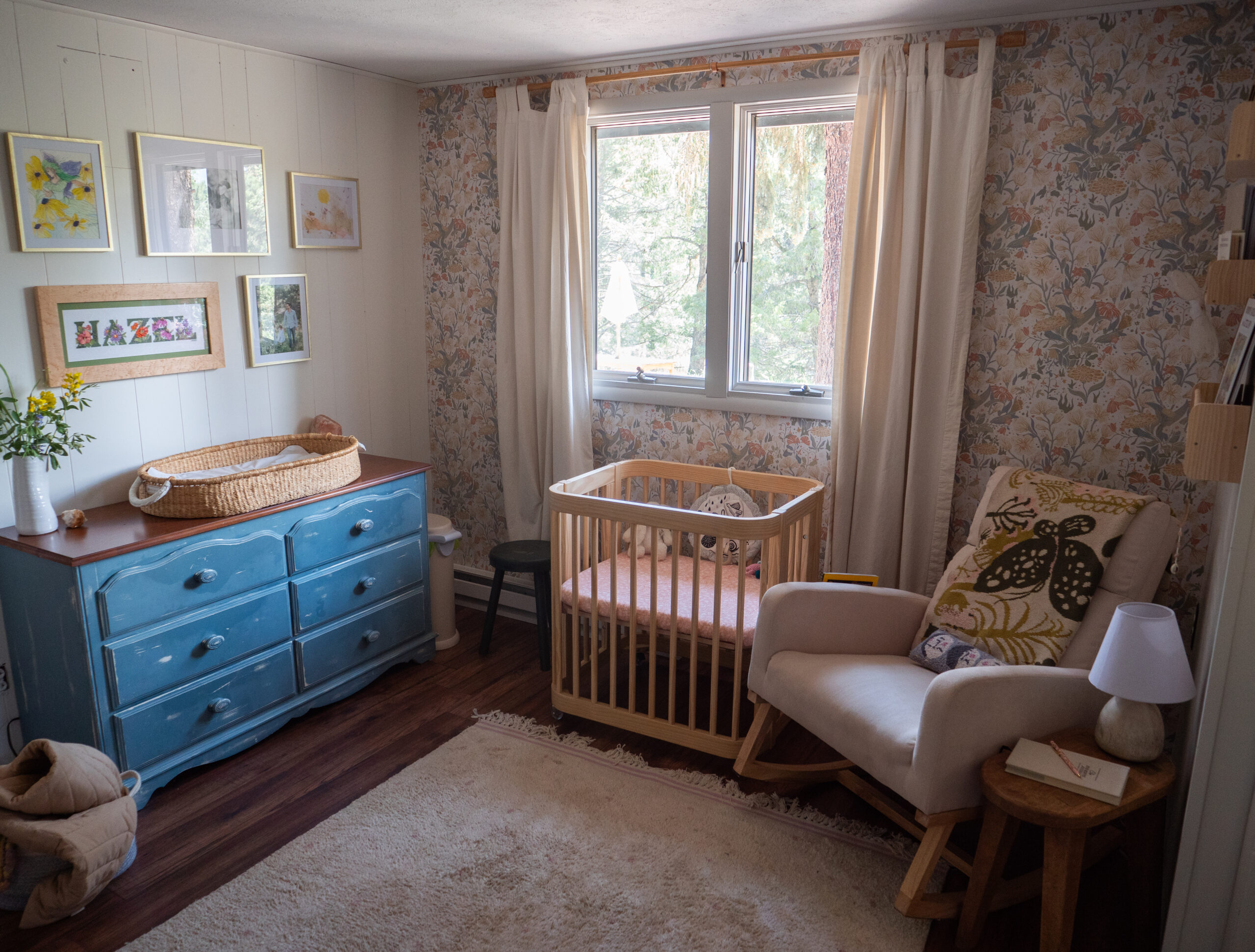 A cozy nursery with a blue dresser, changing basket, crib, armchair with pillows, side table, lamp, and framed art on floral wallpapered walls near a window.