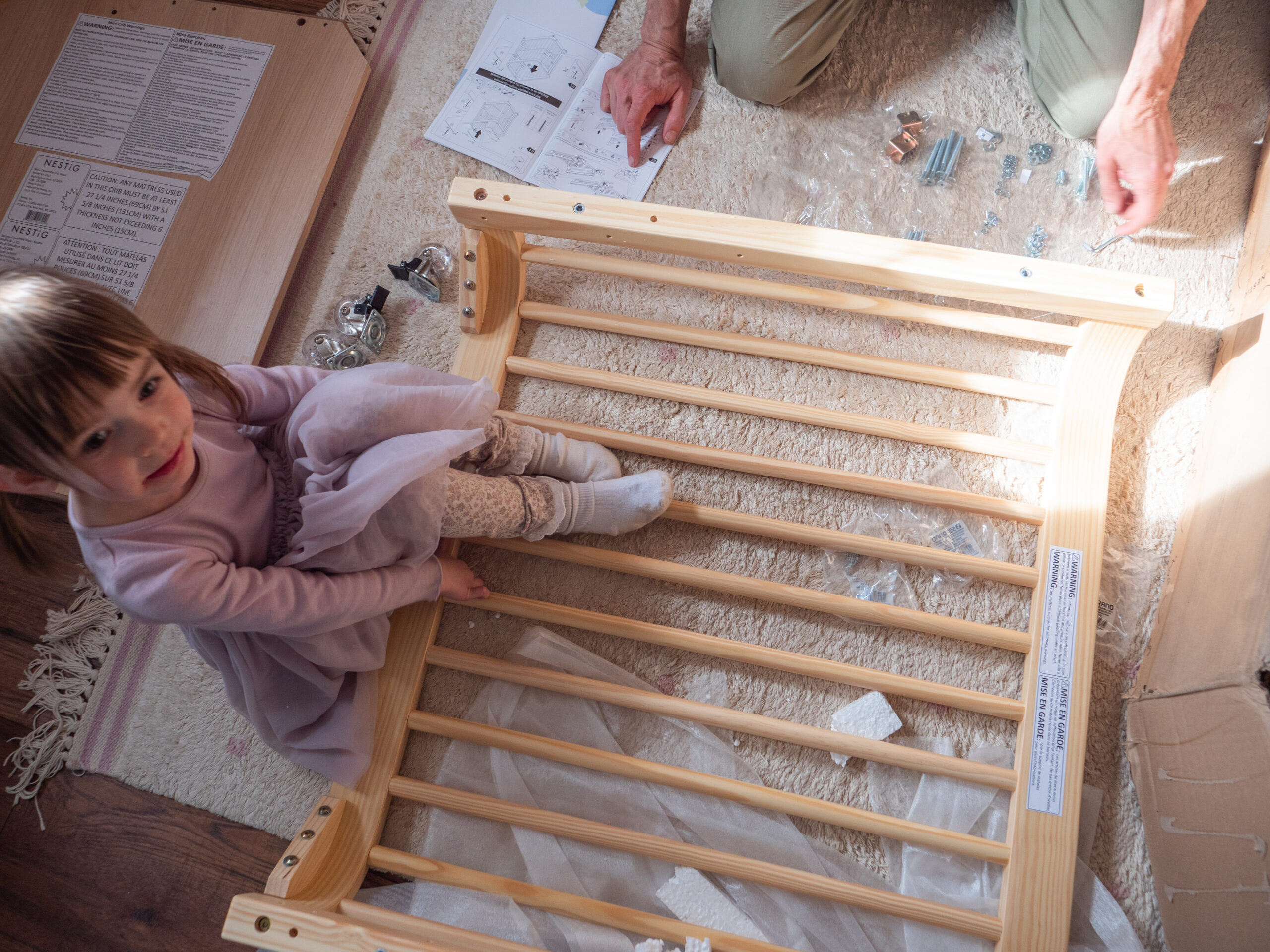 A young girl sits on a wooden crib frame while an adult assembles it on the floor, surrounded by screws, instructions, and other parts.