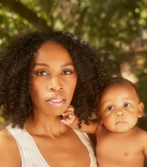 A woman with curly hair holds a baby in her arms outdoors, both looking directly at the camera with a blurred green background.