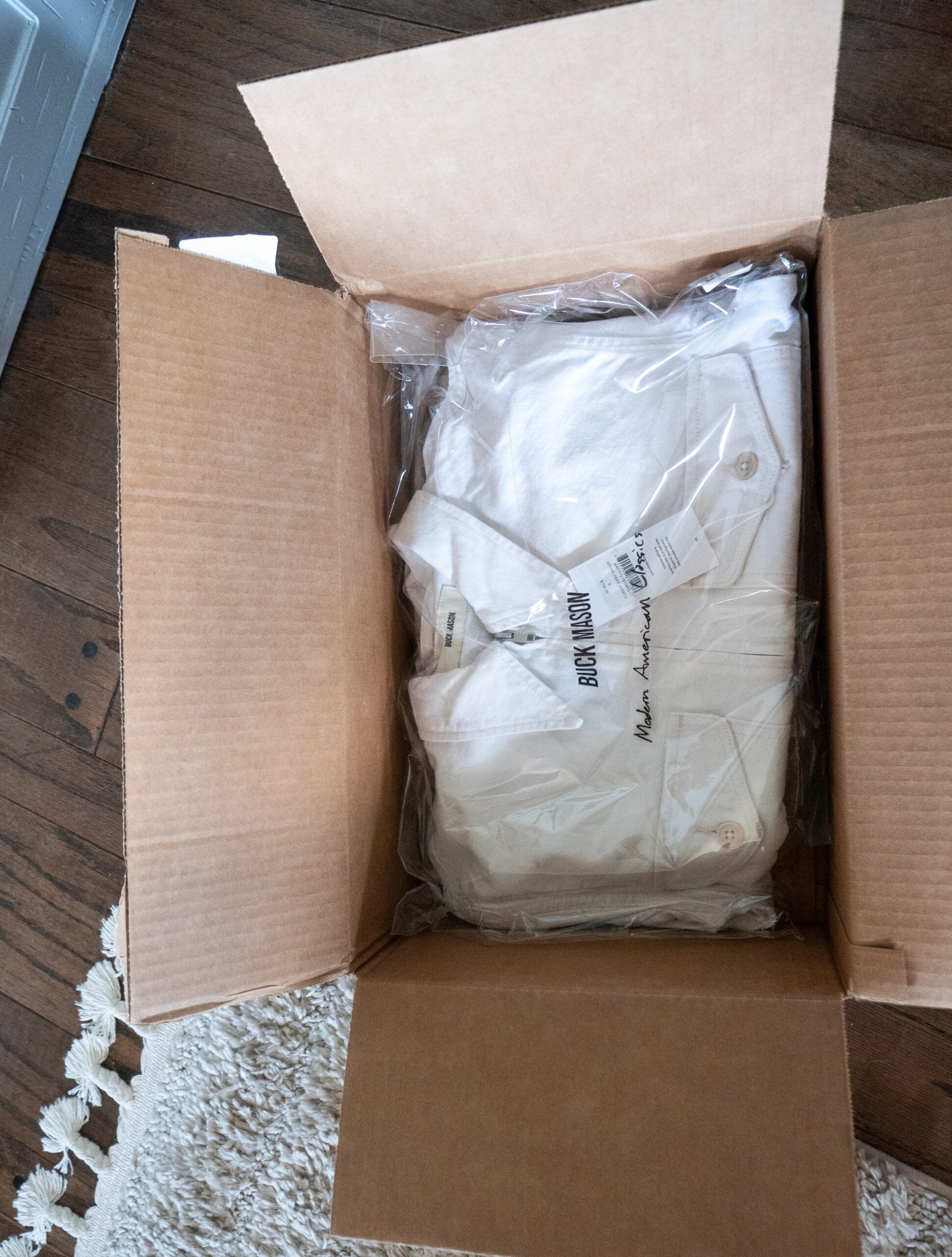 An open cardboard box containing a neatly folded white shirt in plastic packaging sits on a wooden floor next to a beige rug.