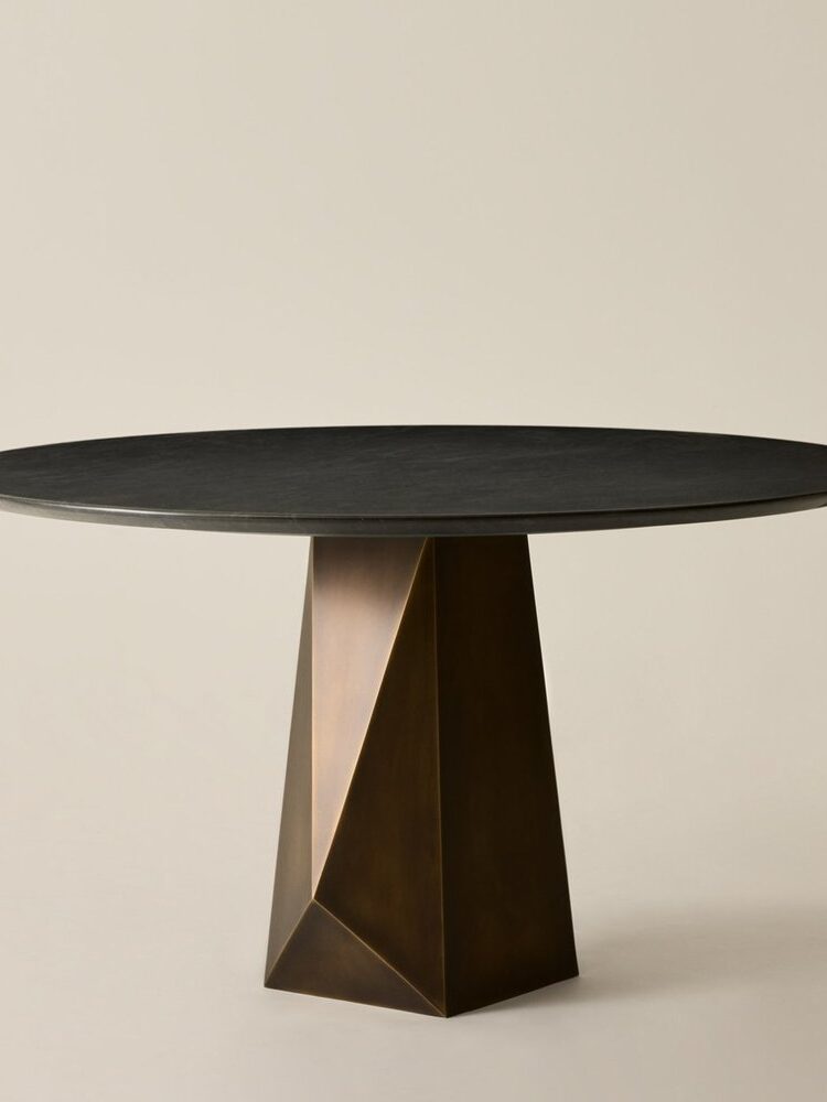A round black table with a geometric, bronze-colored base is set against a plain beige background.