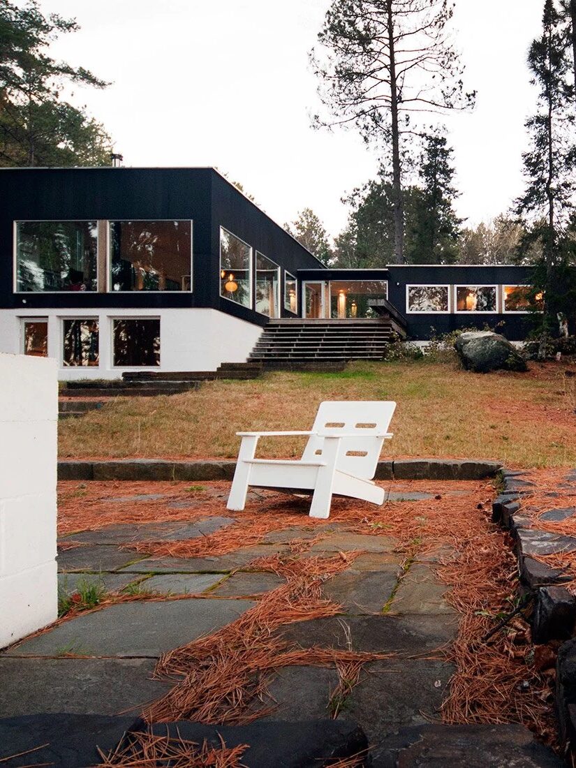 Modern two-story house with large windows surrounded by tall trees, stone patio in foreground, and a white outdoor chair on pine needle-covered ground.