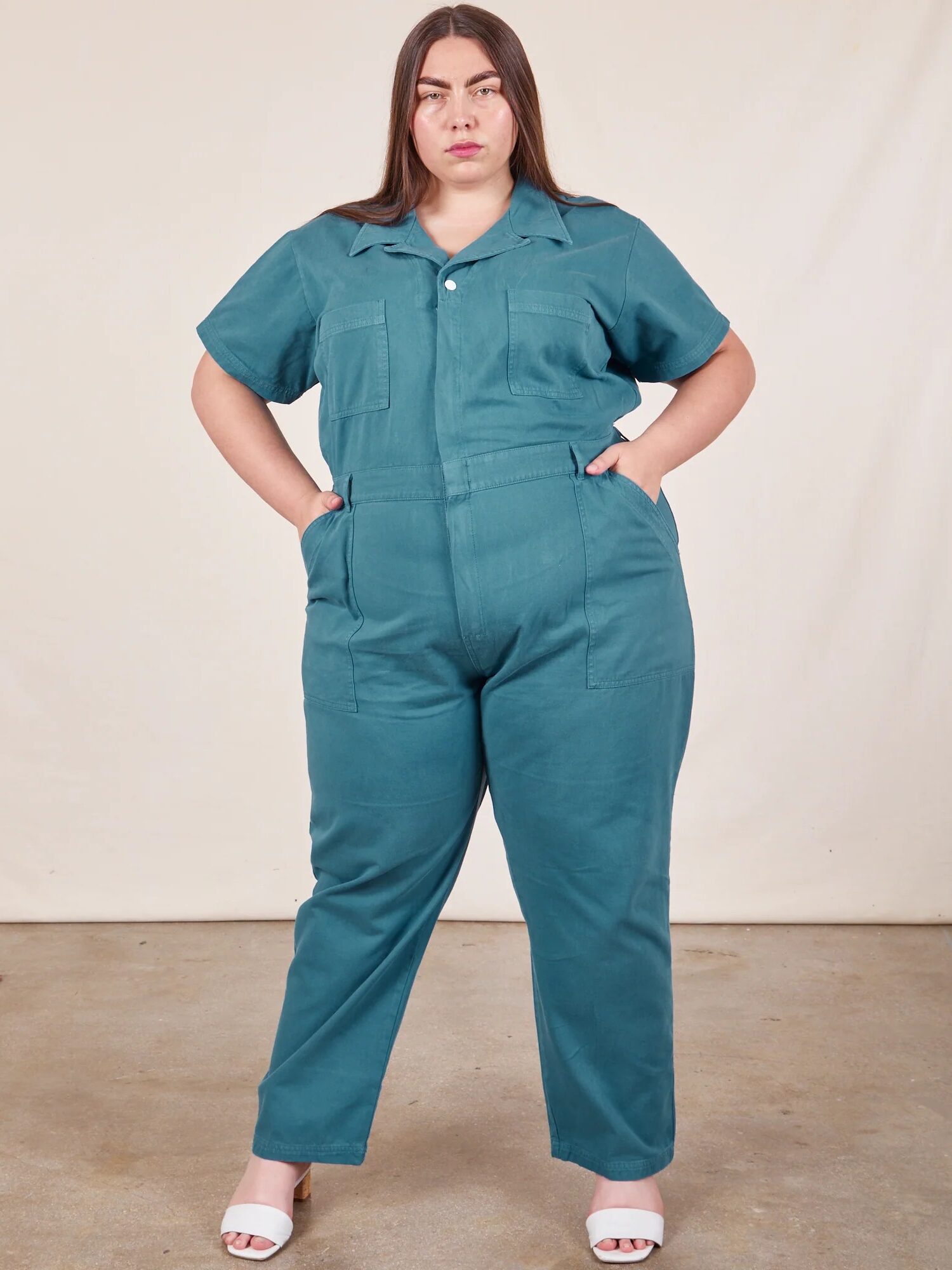 A woman stands indoors wearing a teal short-sleeve jumpsuit with her hands in her pockets, facing the camera against a neutral background.