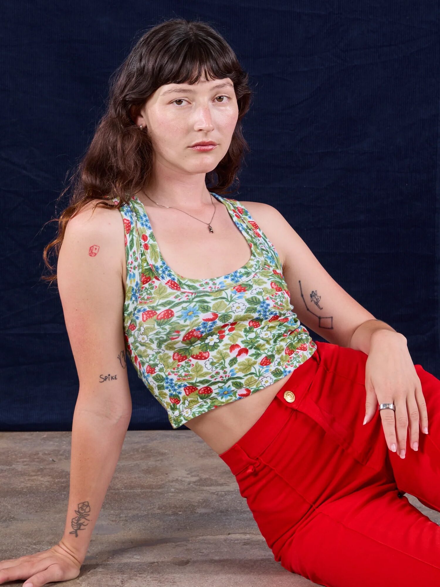 A person with long brown hair sits on the floor, wearing a green floral tank top and red pants, against a dark blue backdrop.