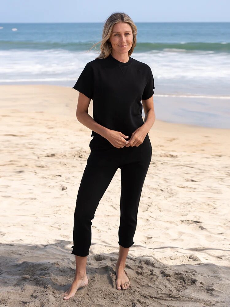 A woman wearing a black t-shirt and pants stands barefoot on a sandy beach with the ocean and waves in the background.